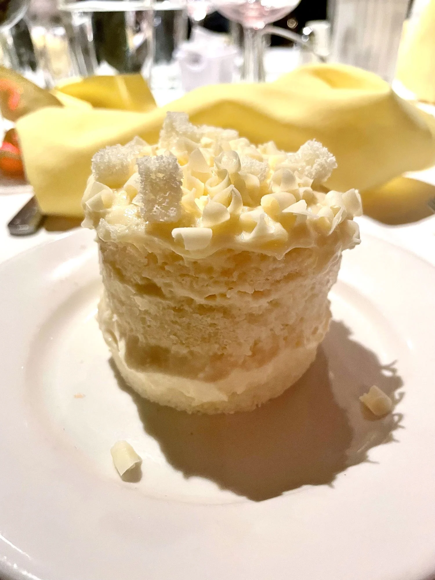 A small white dessert topped with white chocolate shavings, served on a white plate. In the background, a yellow napkin and wine glasses are visible.