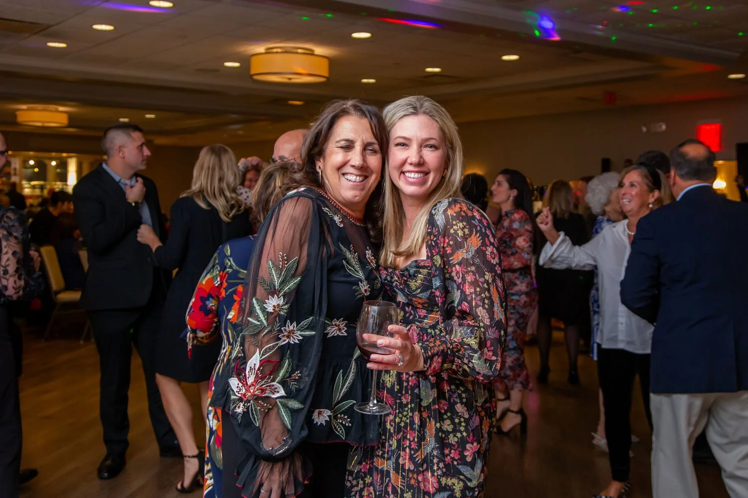 Two women smiling and dancing at a social gathering or party, one holding a glass of red wine, with multiple people in the background socializing and enjoying the event in a large indoor venue.