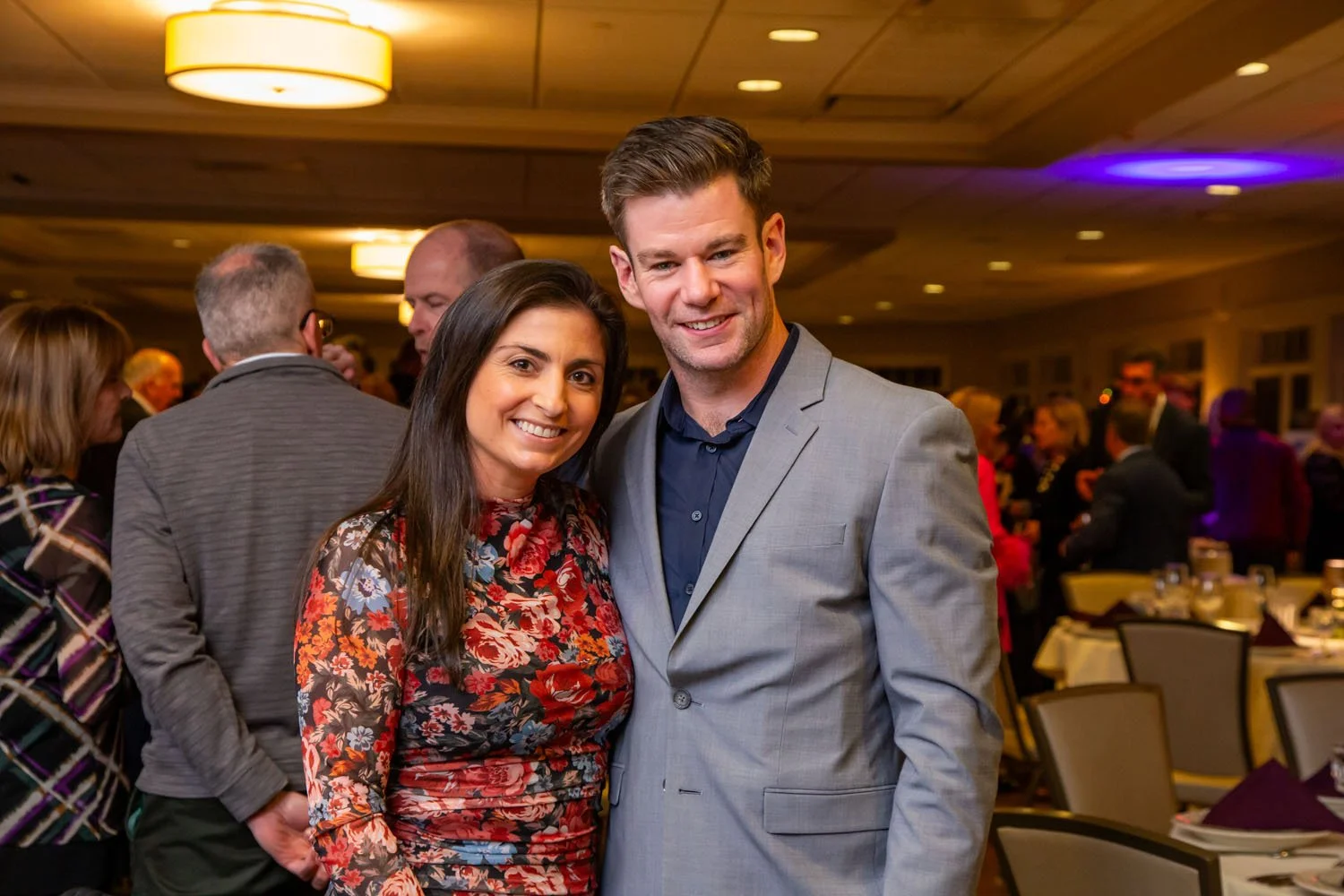 A woman and a man smiling at an indoor social event, with other people in the background, some talking and some standing near tables with purple napkins and dinnerware.