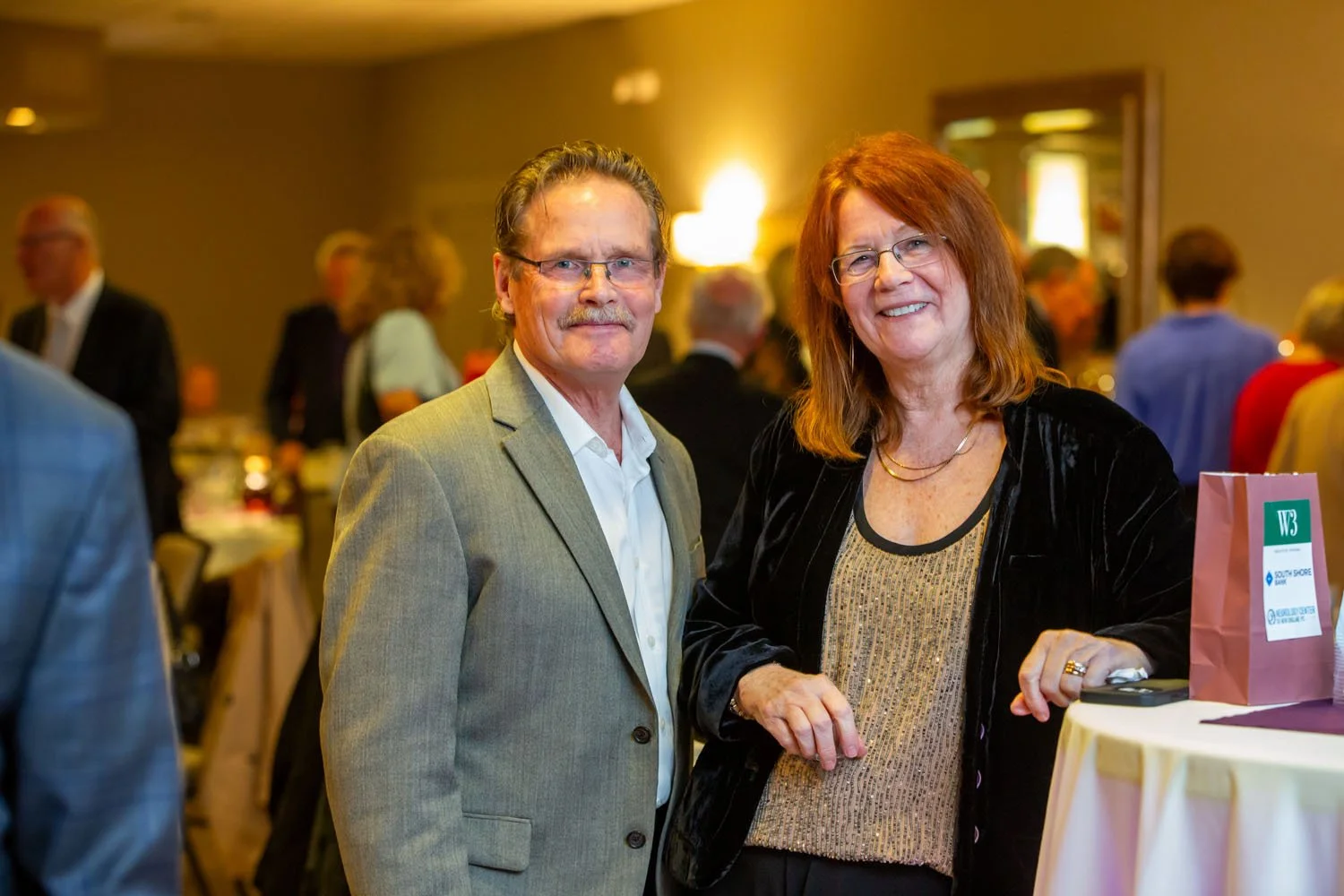 A man in a gray suit and a woman with red hair wearing a black jacket and gold top standing together at a social gathering indoors, smiling at the camera.