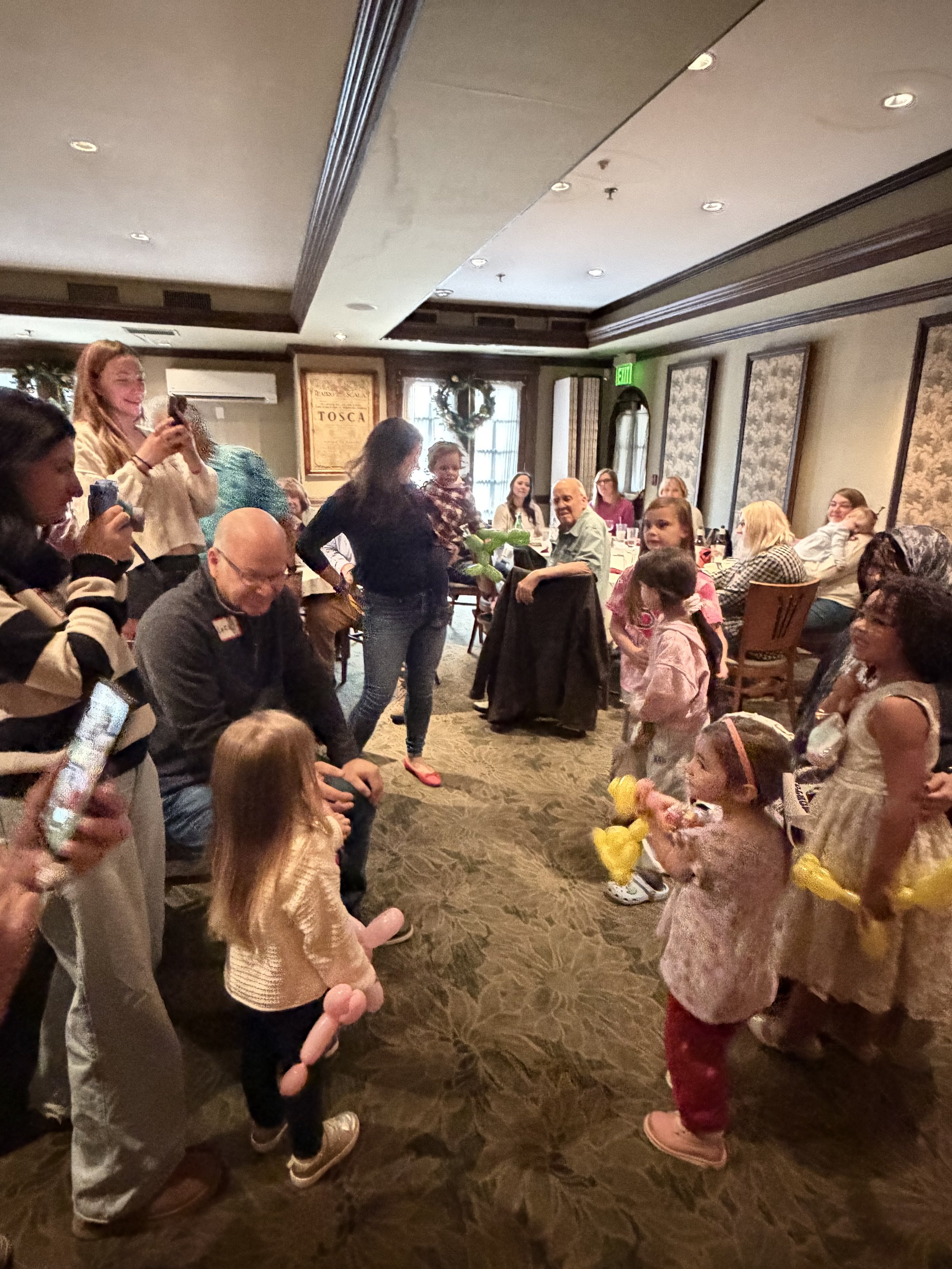 Children and adults gathered in a decorated restaurant for a celebration, with some children holding yellow toys or balloons, and some adults taking photos and watching the children.