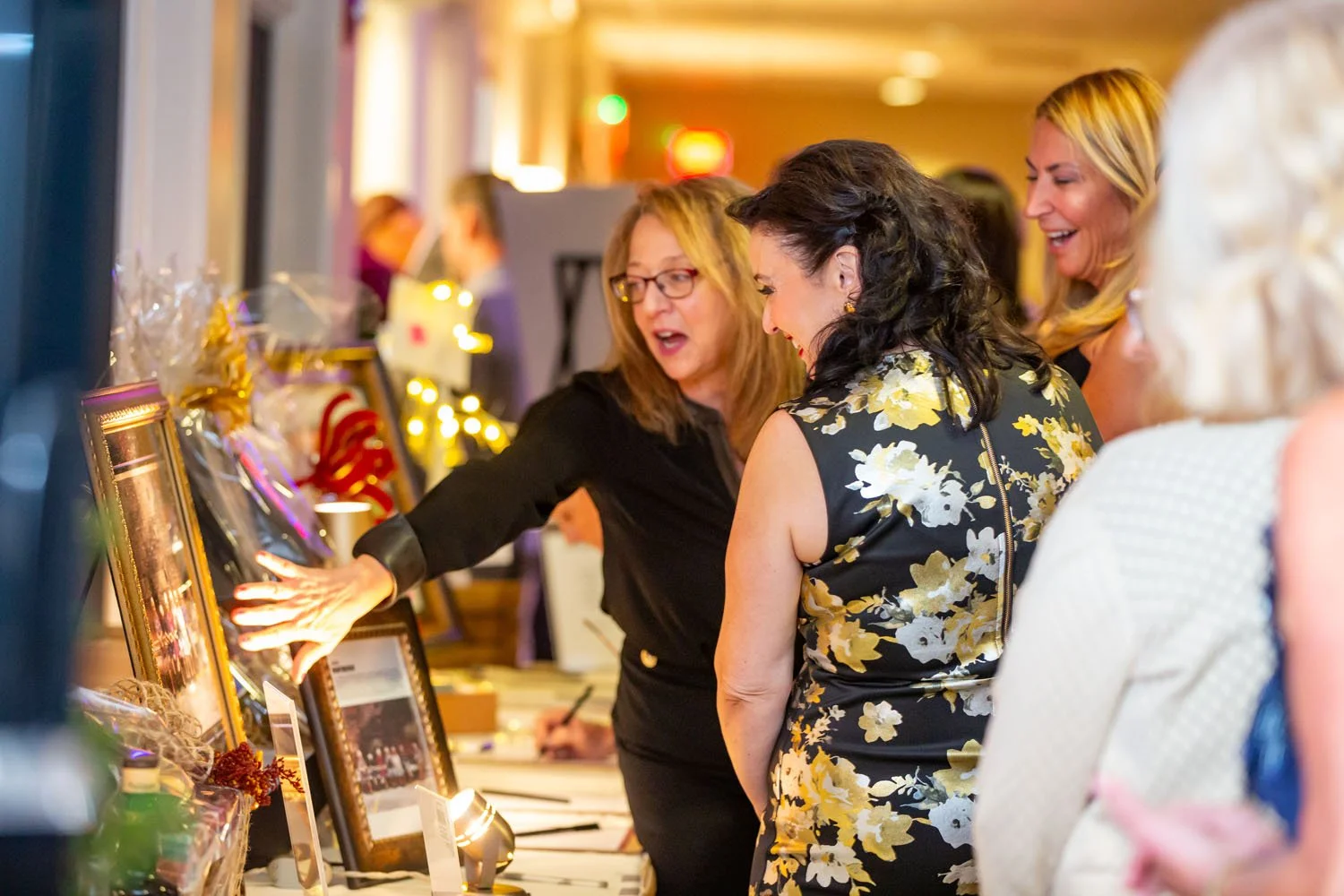 Women browsing art and gift items at a market or auction, with a woman in black pointing at framed pictures on a table, all smiling and engaging
