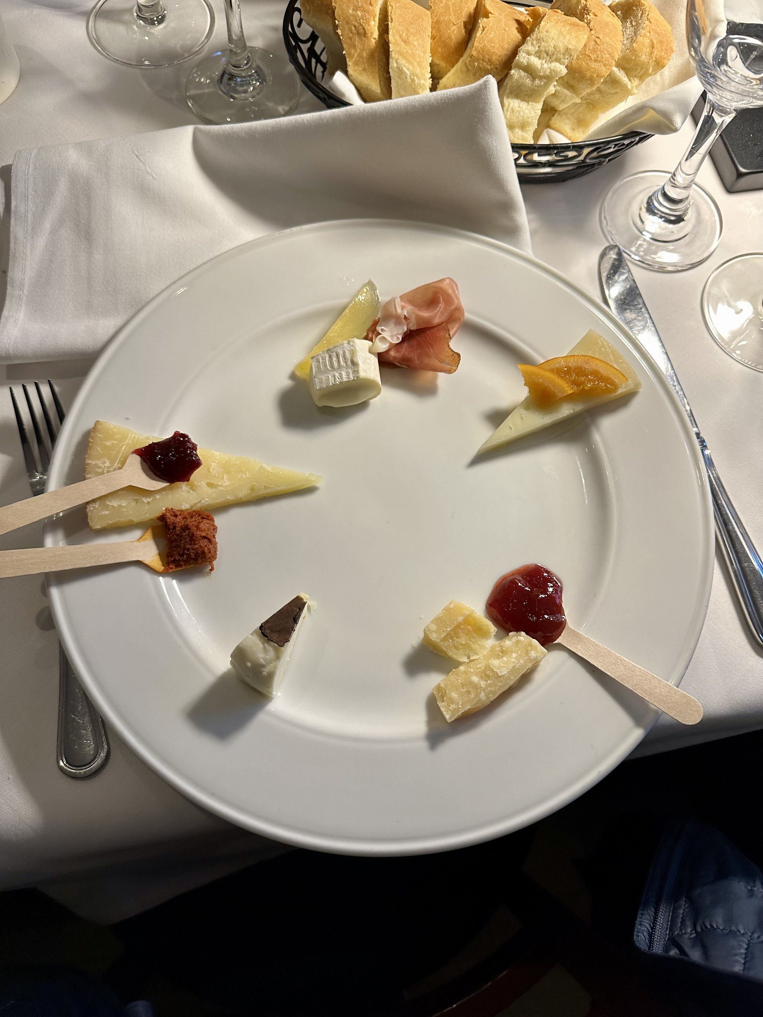 A white plate with a selection of cheeses, meats, and spreads. The cheeses are arranged in a circle, some with toppings like jams. A basket of bread is in the background.