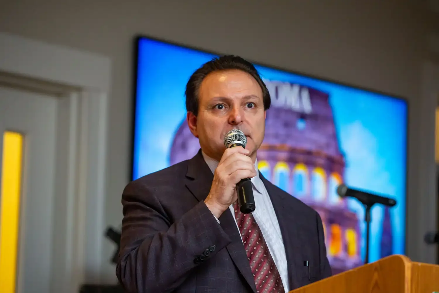 A man in a suit speaking into a microphone at a podium, with a blurred background showing a large screen displaying an image of the Roman Coliseum 