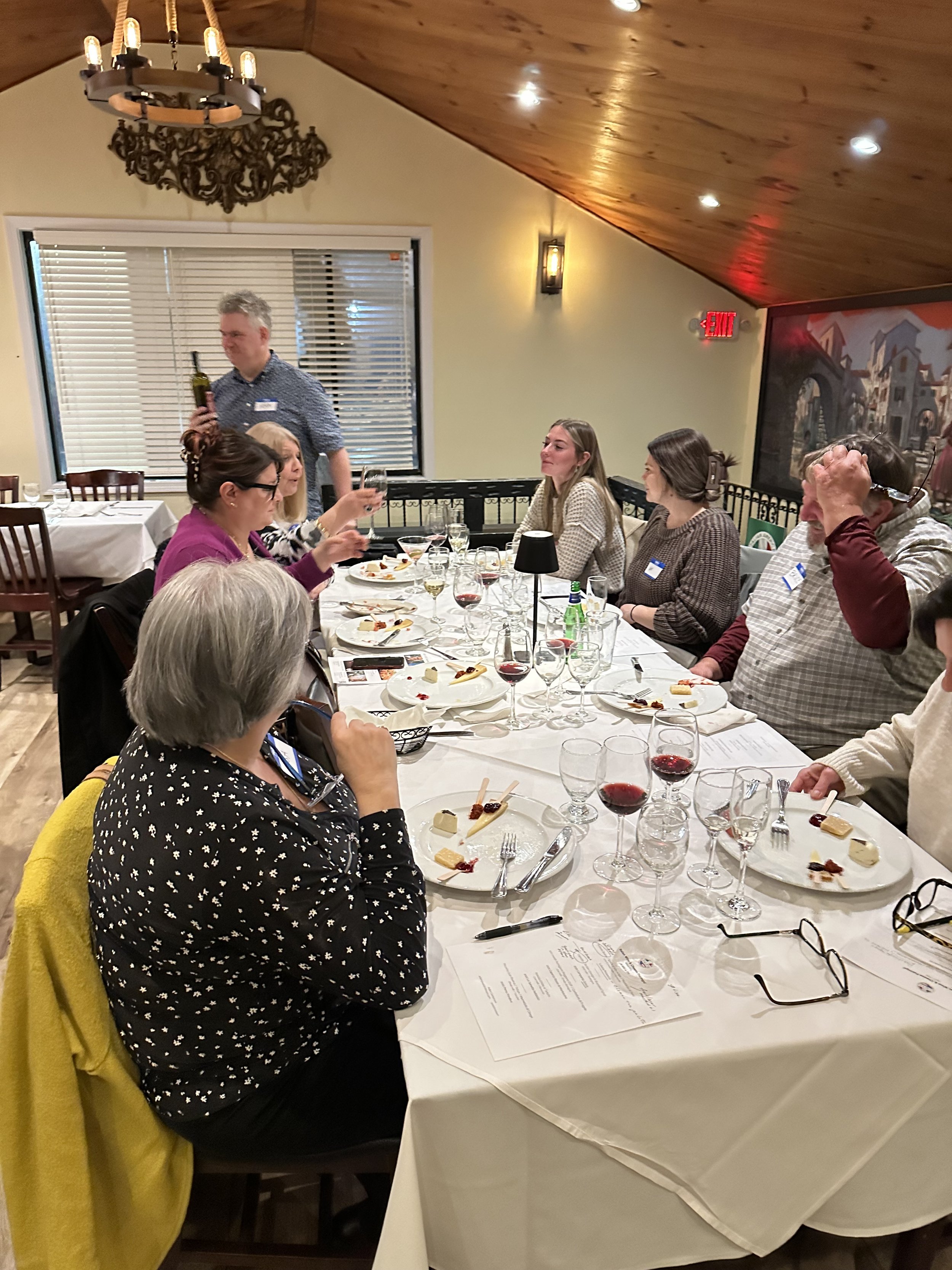 People seated at a restaurant table enjoying a wine and cheese tasting event, with wine glasses and cheese plates on a white tablecloth, under a wooden ceiling with a chandelier.