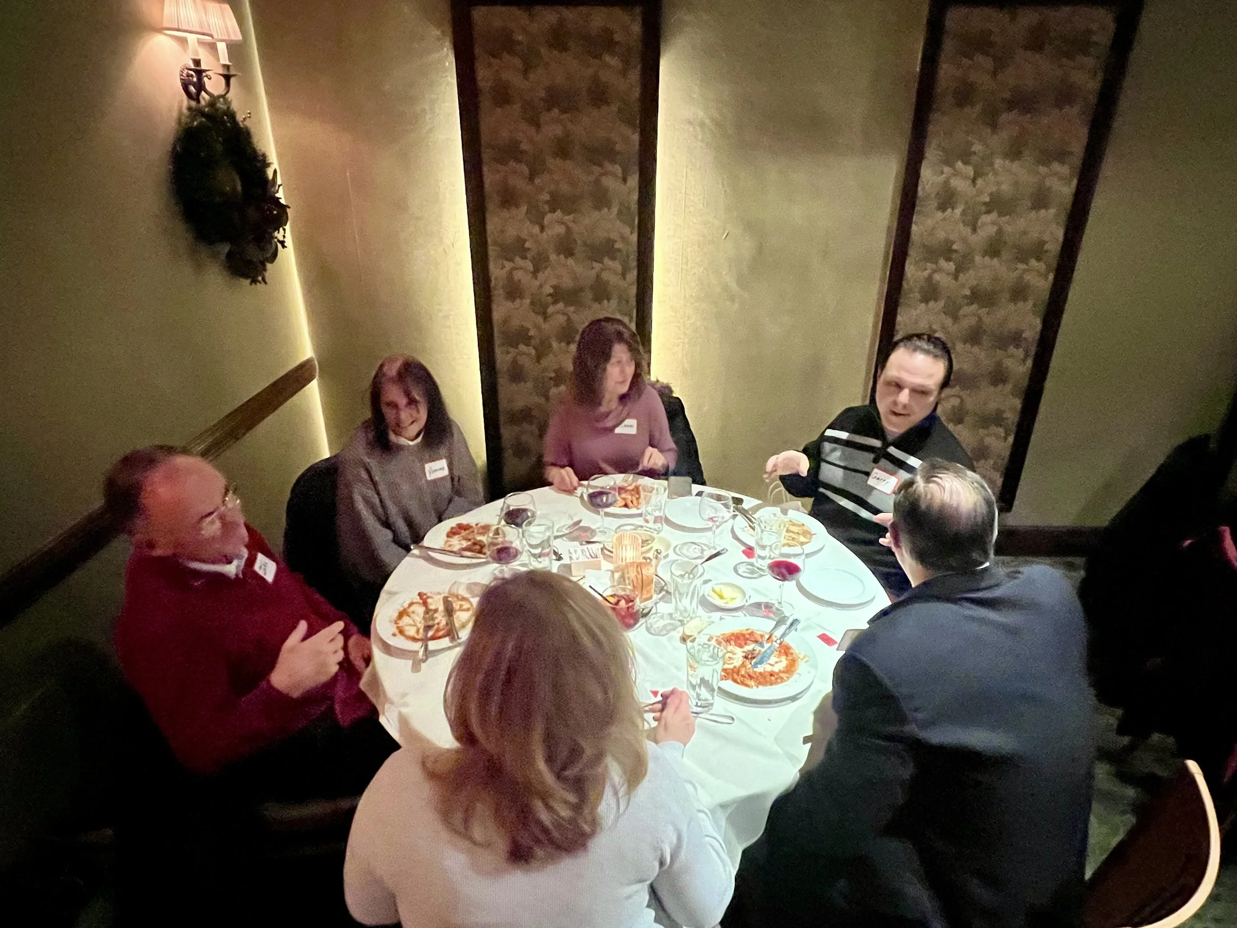 People dining at a round table in a restaurant, eating pizza and drinks, with five individuals visible.