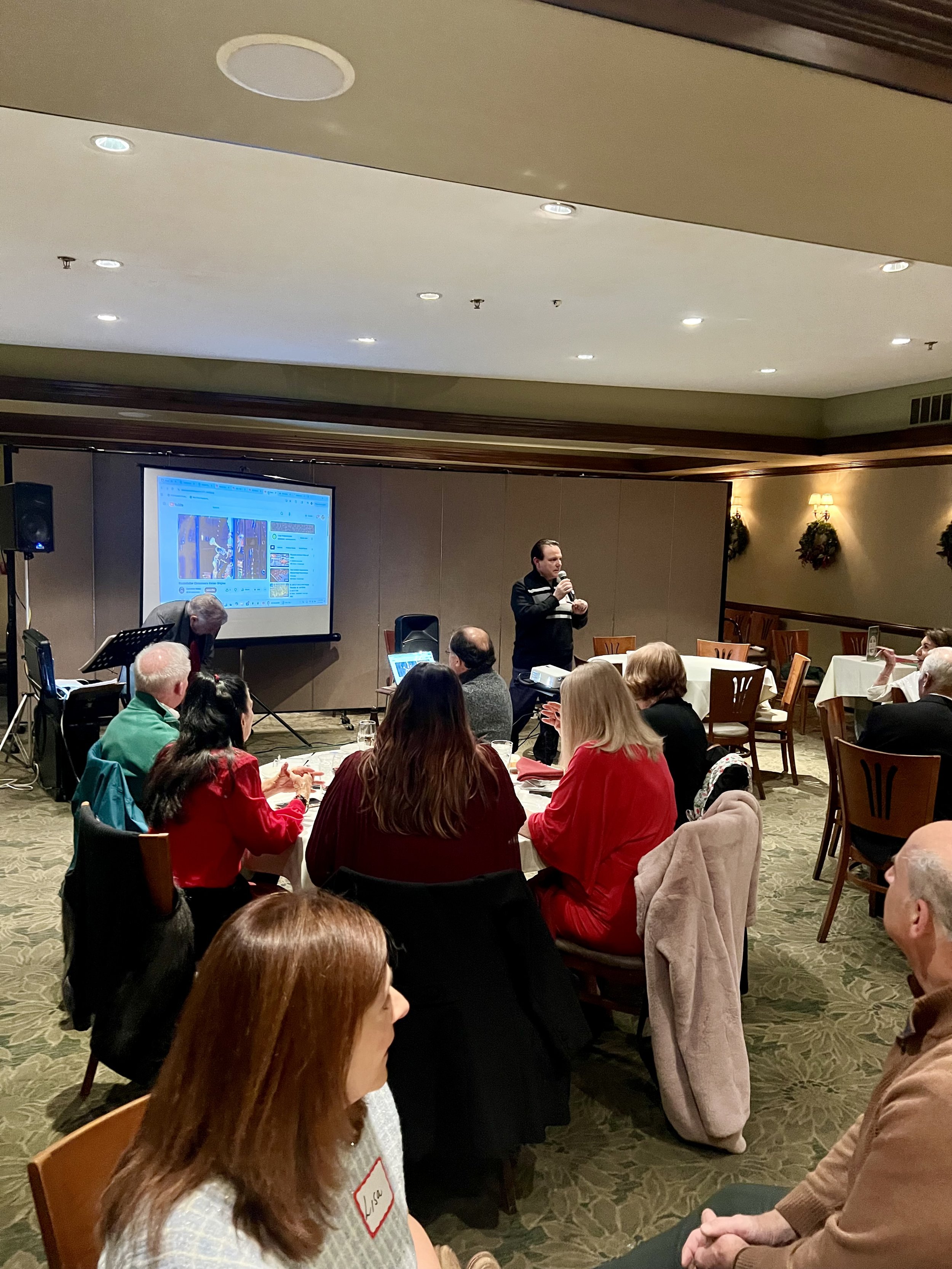 A man giving a presentation to an audience in a banquet room, with a large screen displaying a social media page behind him.
