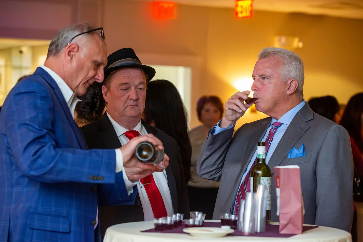 Three men at a social event, two are examining a small bottle, and one is drinking from a glass, with a table holding drinks and snacks in front of them.