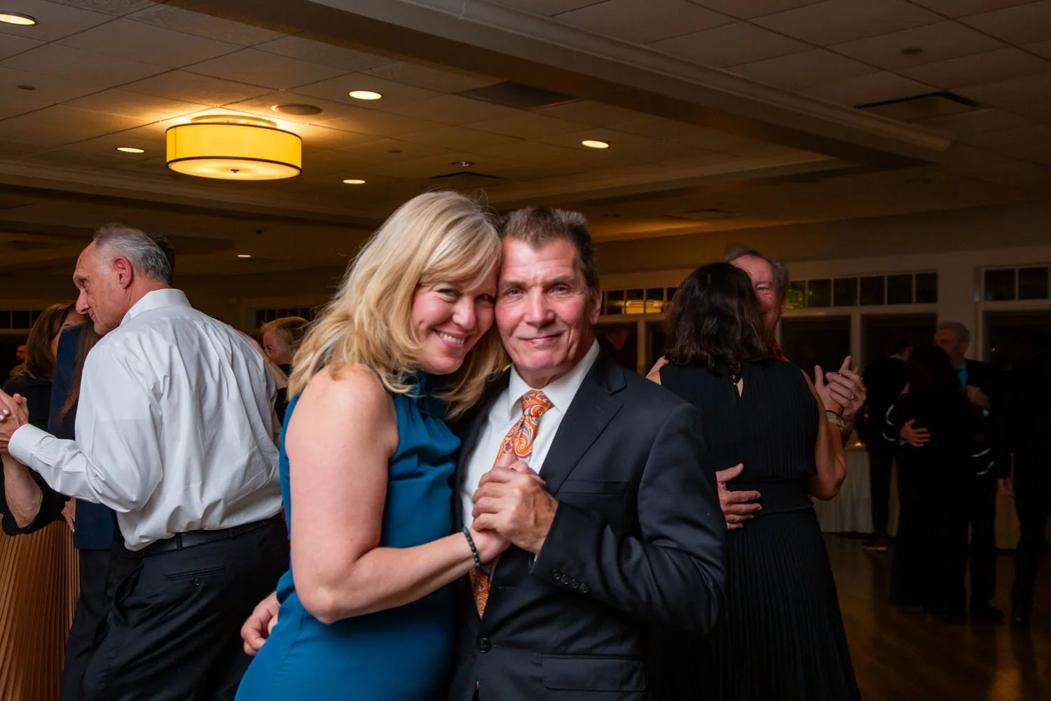 A couple dancing together at a social event, with other couples dancing in the background inside a banquet hall.