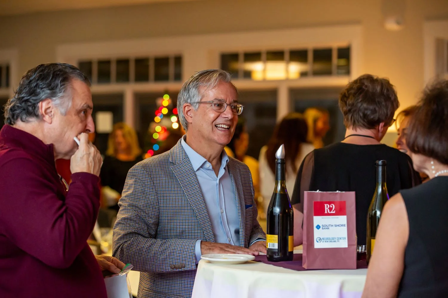 People socializing at a festive gathering, with a Christmas tree in the background, standing around a cocktail table with drinks and a pink bag.