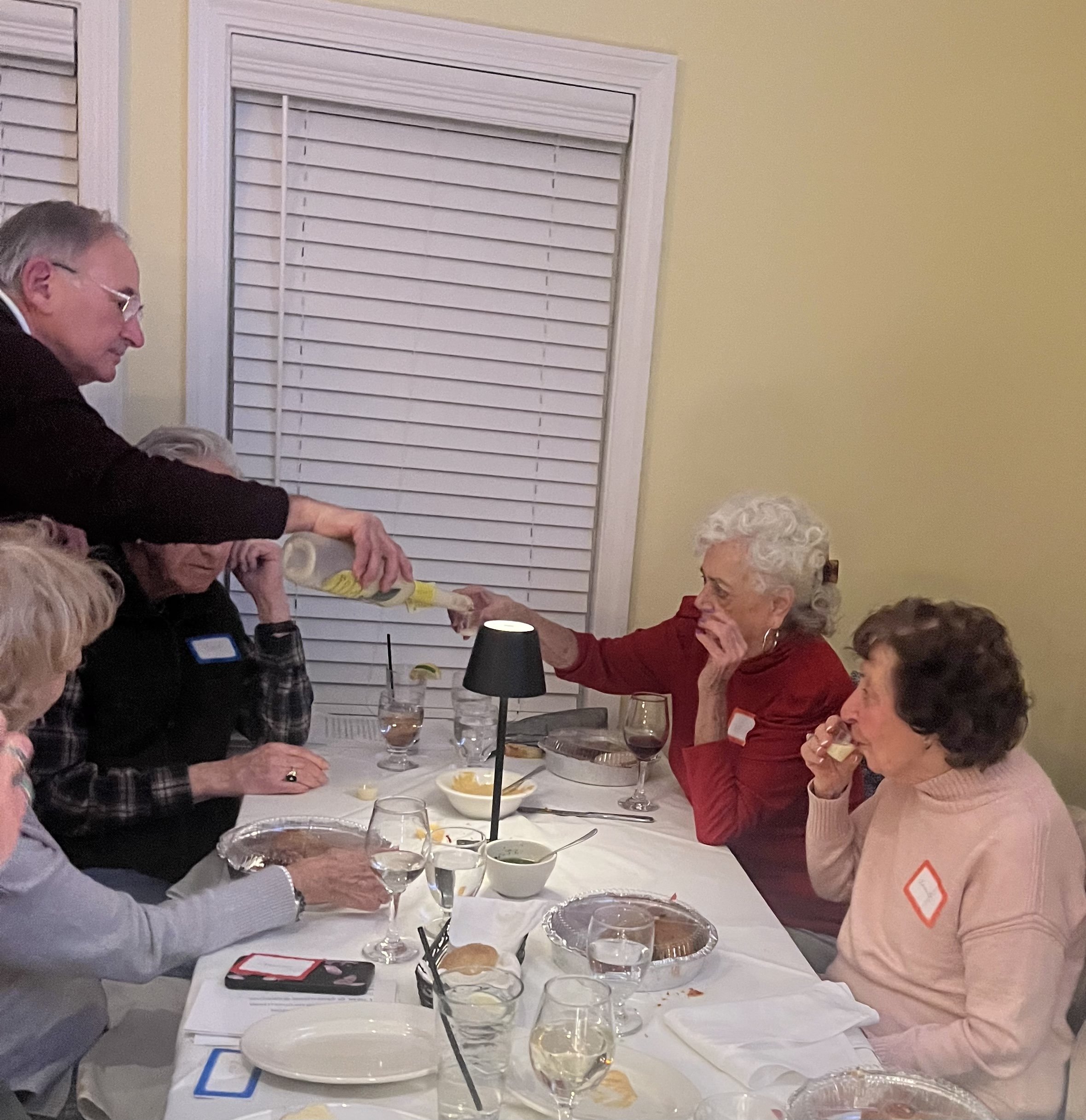 A group of older adults sitting at a dining table, engaged in conversation. One person is pouring a drink, while others have food and drinks in front of them. The setting is casual, with plates, glasses, and a small lamp on the table. The room has li