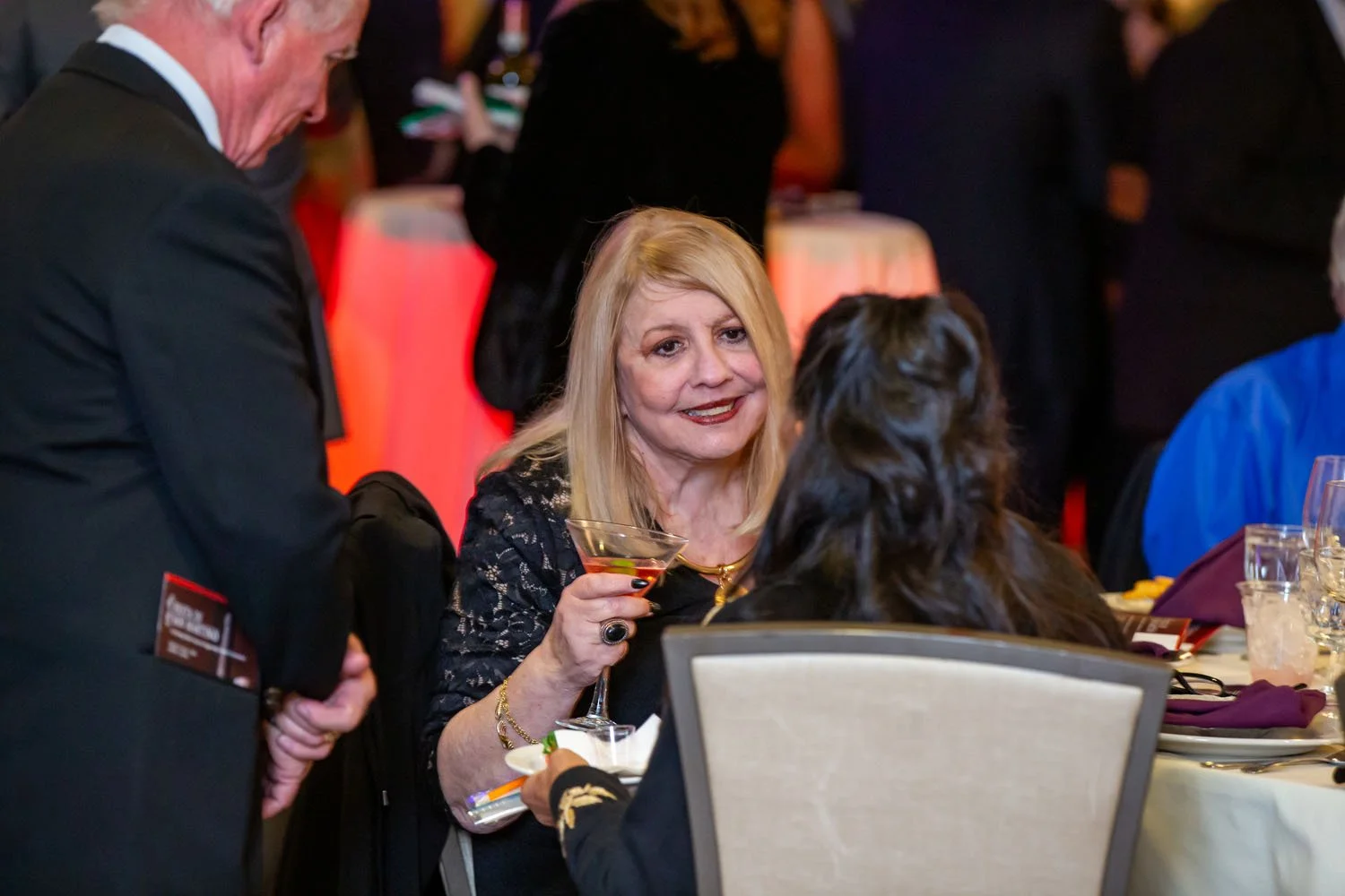 A woman with blonde hair holding a cocktail and smiling at a social event, with other people in the background.