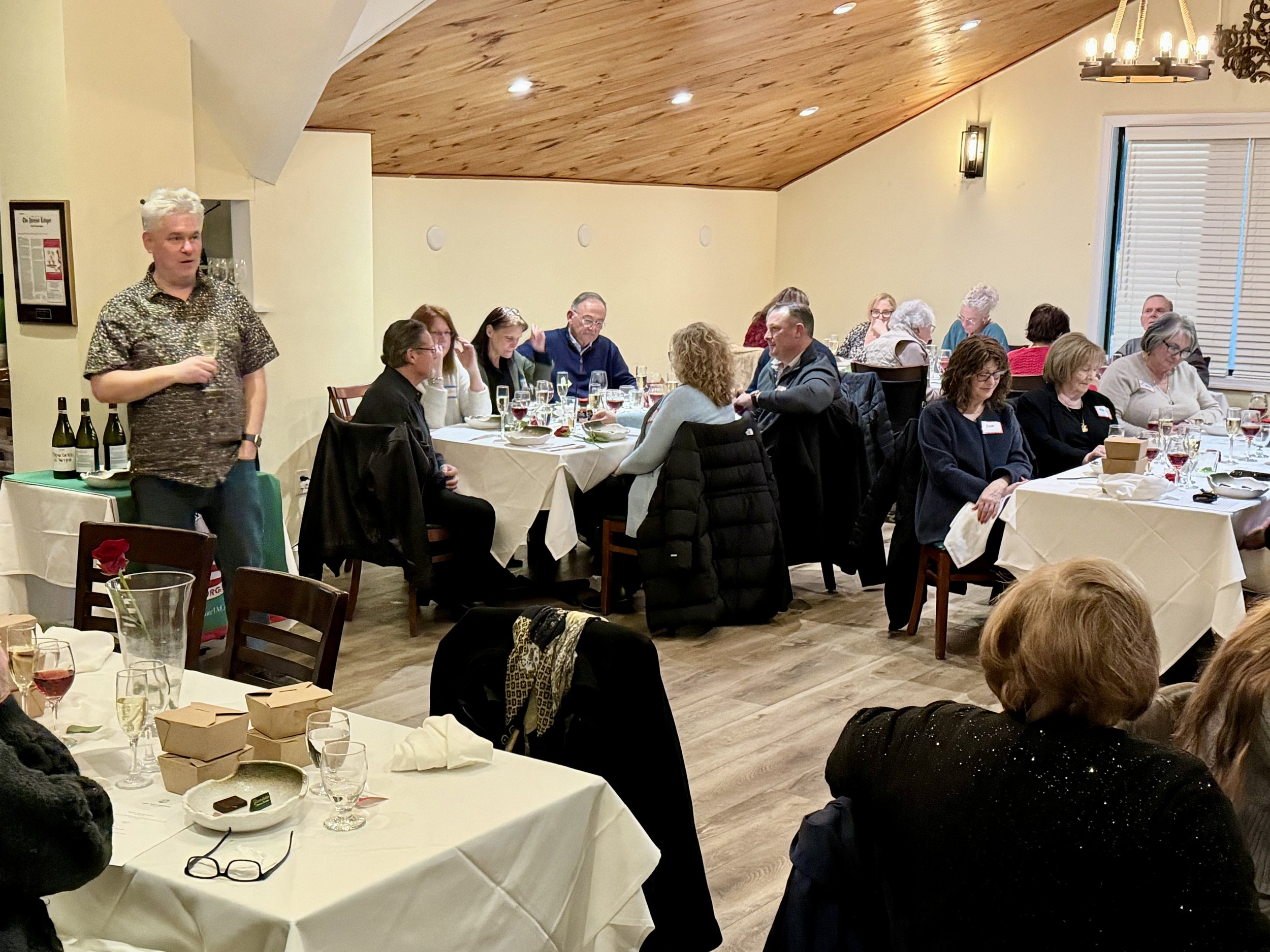 A gathering of people sitting around a banquet table in a restaurant, with one man standing and speaking, while others listen and interact. The room has wood-paneled ceilings, cream walls, and a mix of decorative and functional lighting.