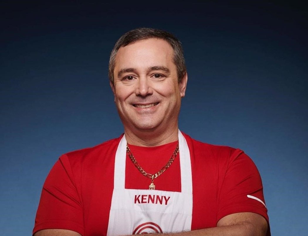 A smiling man wearing a red shirt, white apron with the name 'Kenny', and a gold chain necklace.