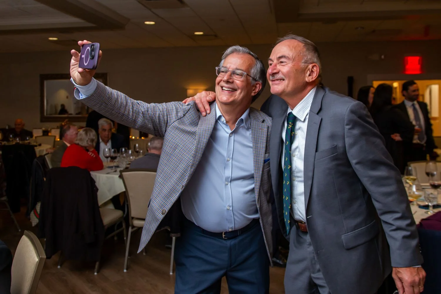 Two middle-aged men in suits taking a selfie together at a formal event, with other guests seated at tables in the background.