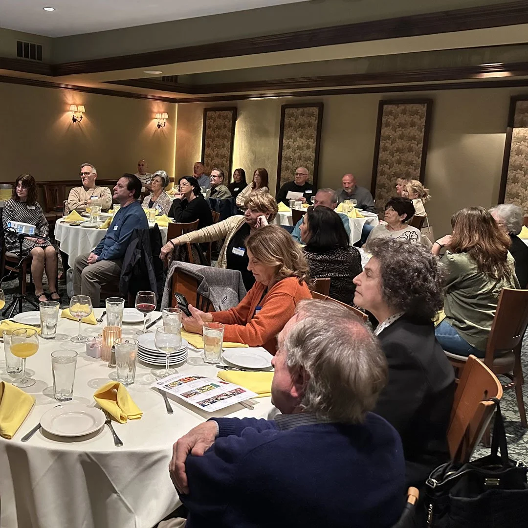People attending a formal event or conference in a banquet hall, sitting at round tables with yellow napkins, glasses, and plates.