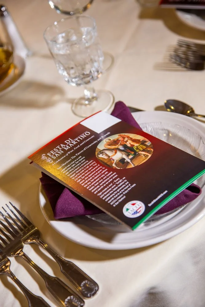 A restaurant table setting with a white plate holding a purple napkin and a pamphlet about San Martin's feast, a water glass with ice, and silverware including a fork and spoon.
