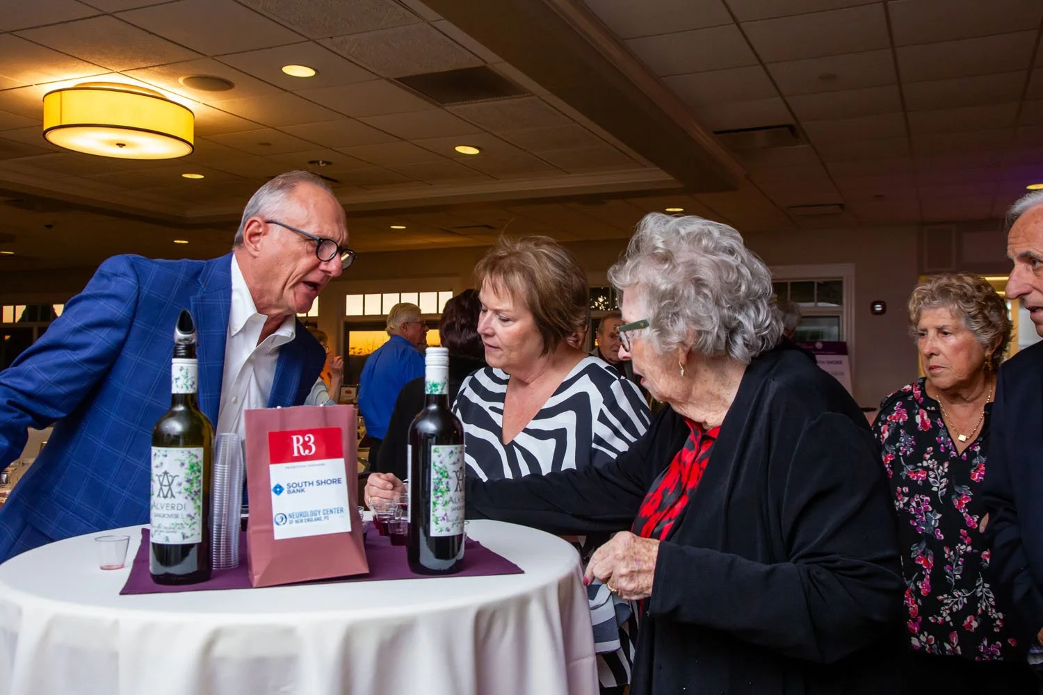 A group of older adults around a table at a social event, with bottles of wine and a pink gift bag on the table, indoors with warm lighting.