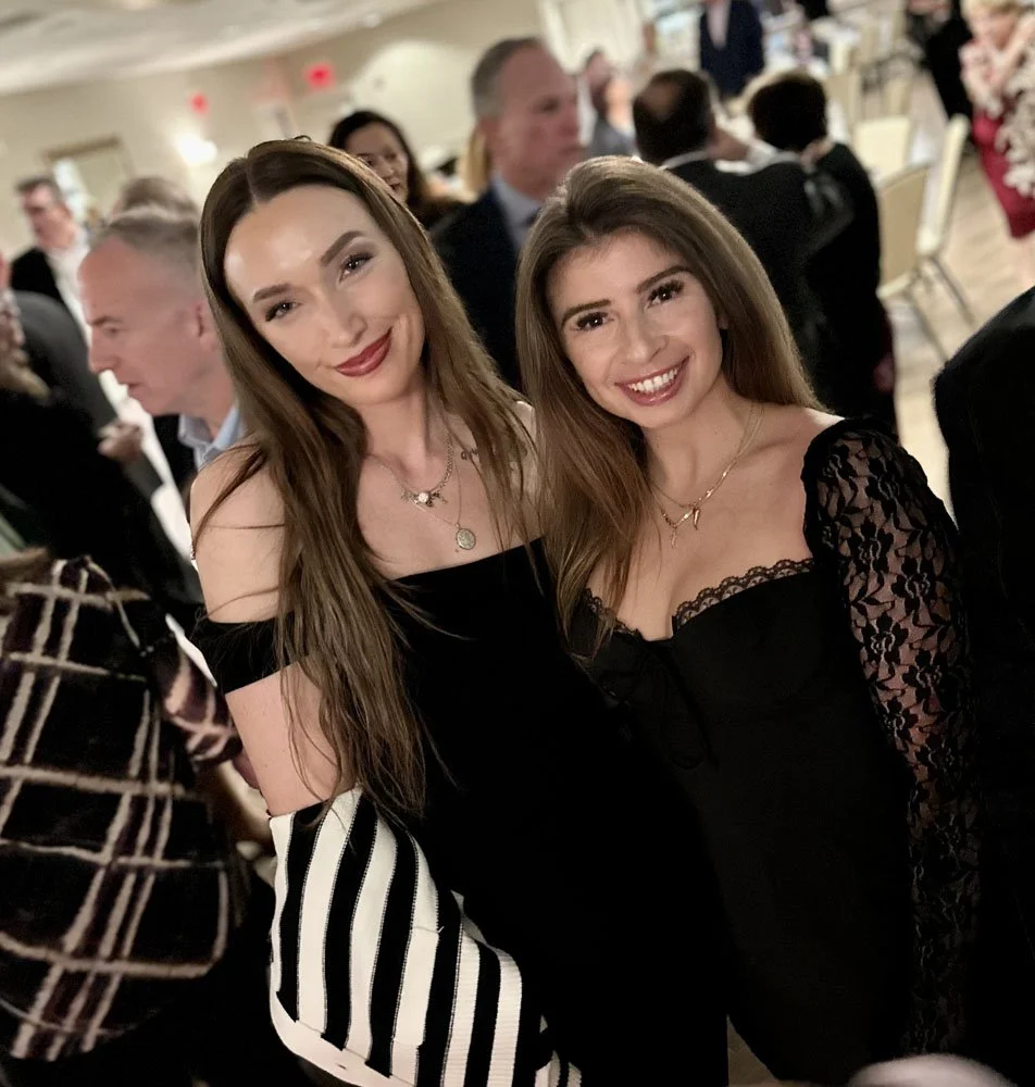 Two women dressed in black at a formal event, smiling for a photo, surrounded by other guests in the background.