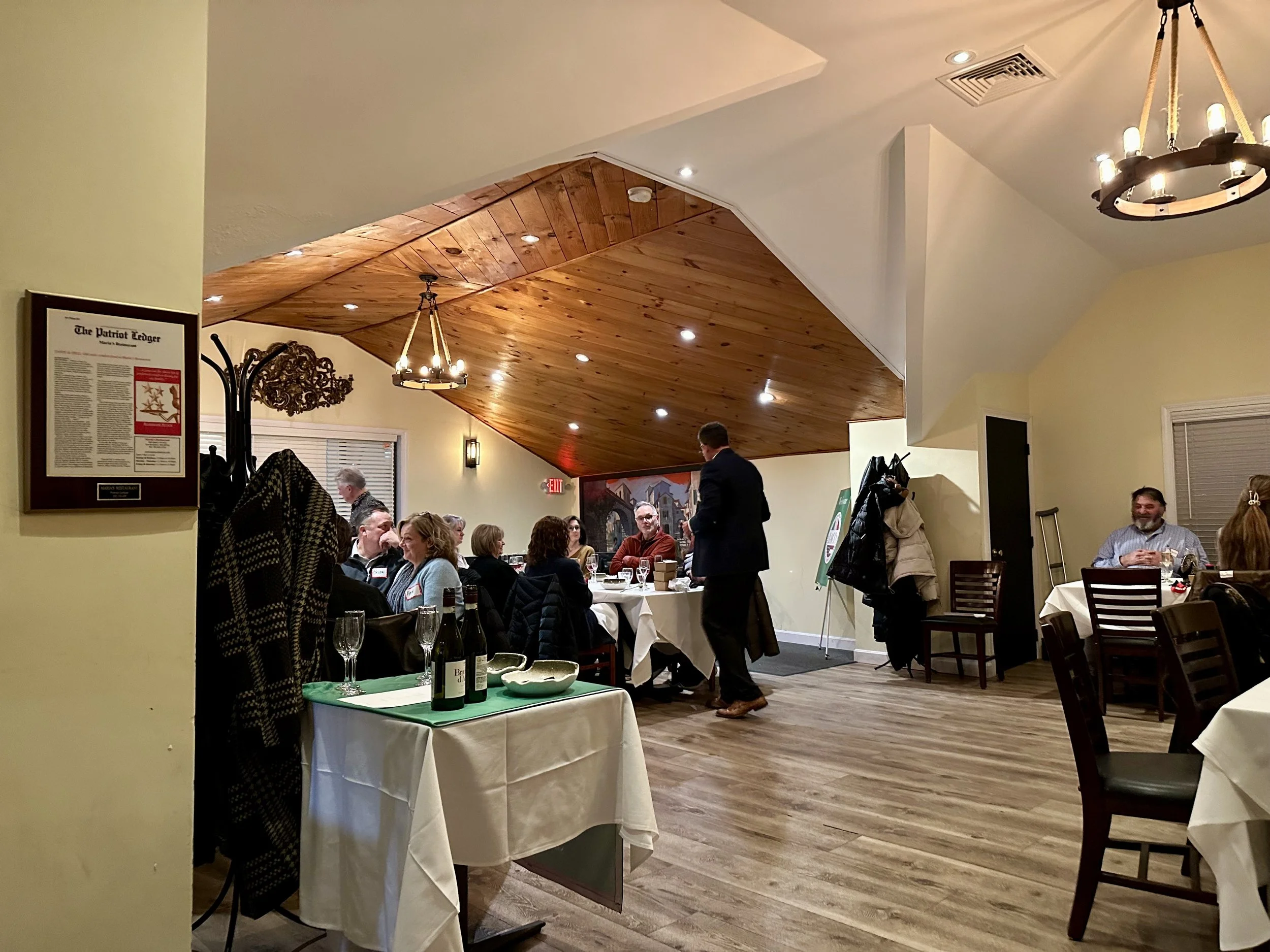 A restaurant dining area with several people seated at tables, some engaged in conversation. The room has wooden and beige walls, a wood-paneled ceiling, and warm lighting fixtures. There is a table near the camera with wine bottles and glasses, and 