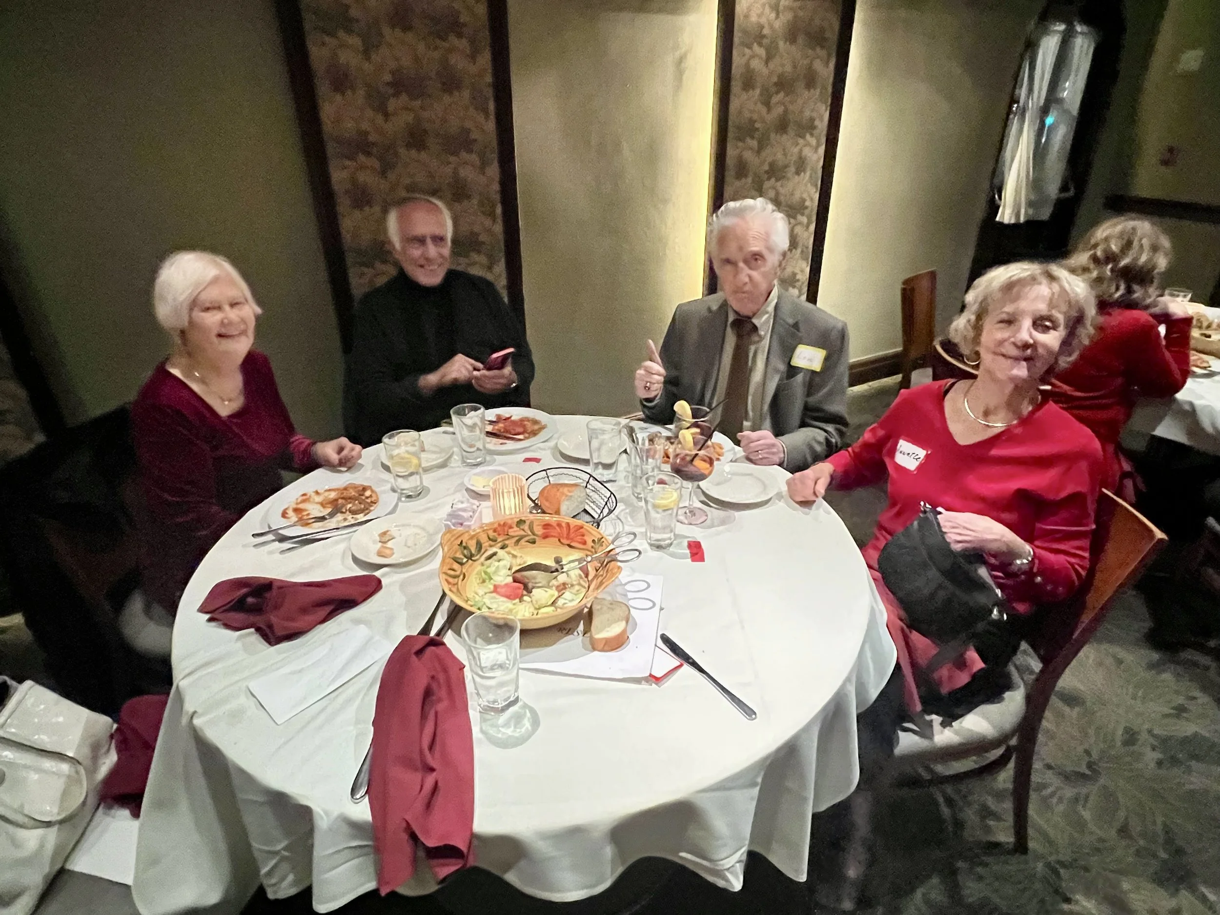 Four elderly people, three women and one man, are sitting at a round dining table with white tablecloth, eating and smiling. The table has plates with food, glasses of water, and a large decorative bowl of salad in the center. The women are dressed i