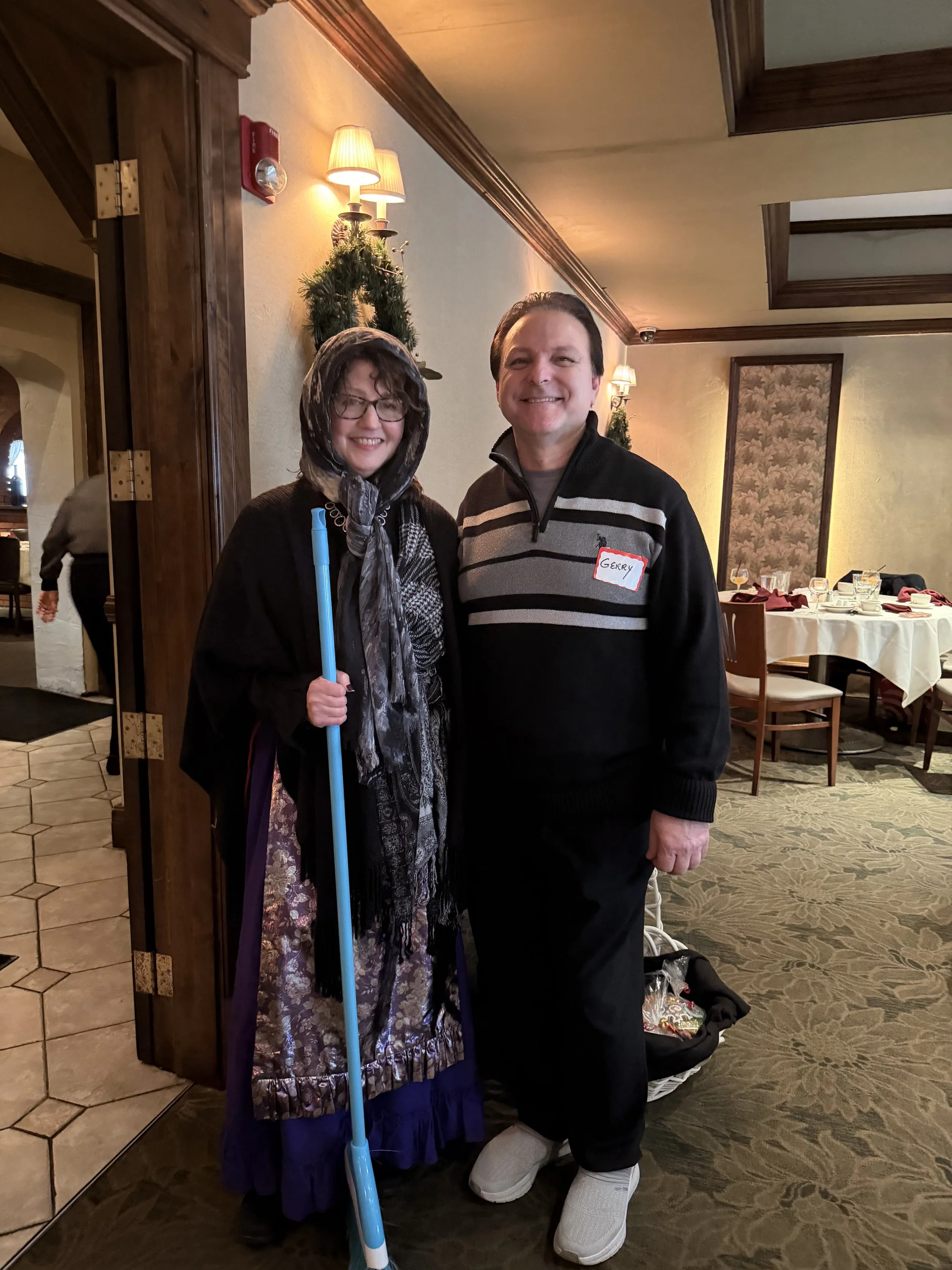 A man and woman standing together in a decorated indoor setting, smiling at the camera. The woman holds a blue cane, wearing glasses and a scarf. The man is wearing a black sweater with gray stripes and has a name tag that says 'Gerry'.