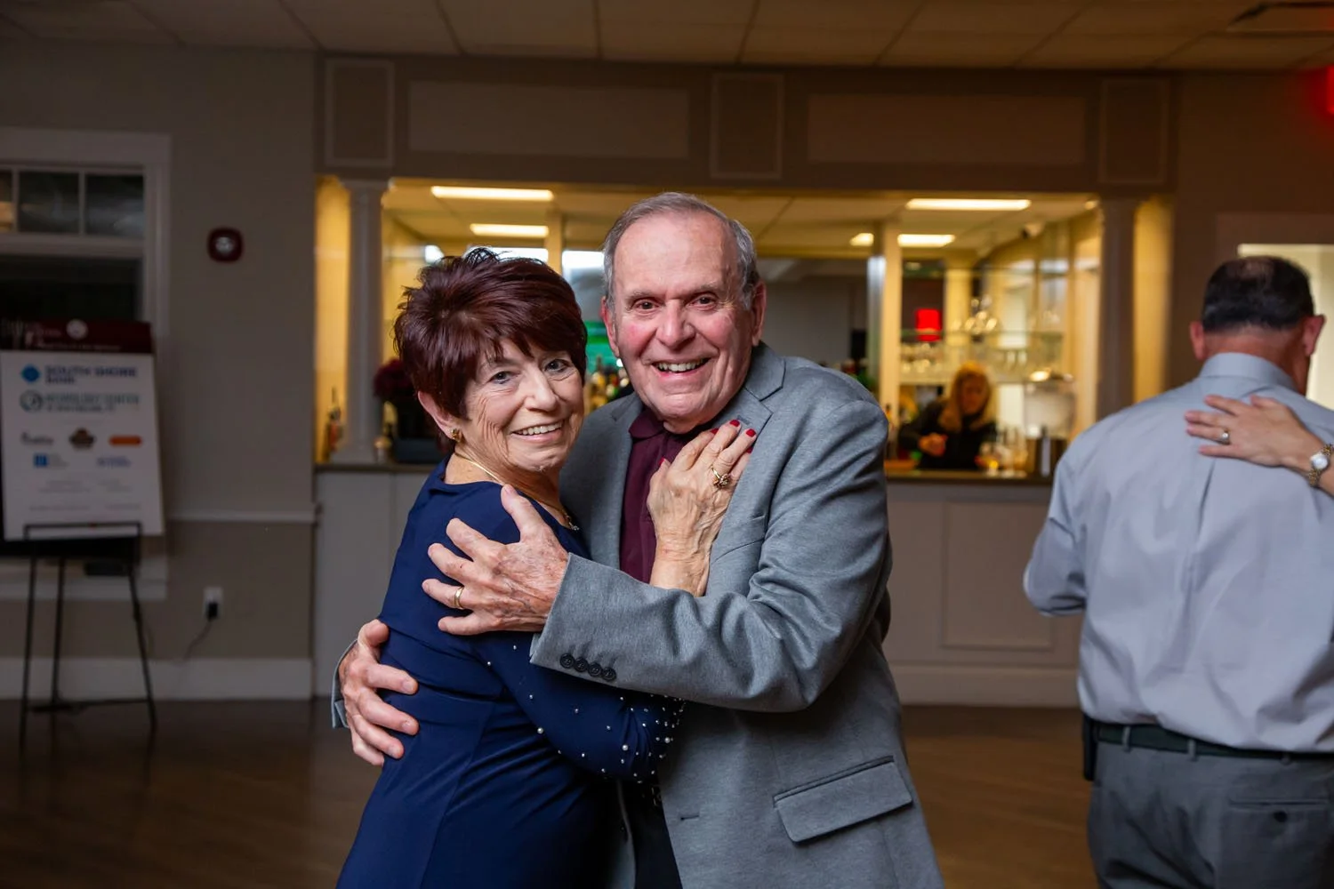 An elderly couple dancing and smiling at a social event in a well-lit indoor venue, with a woman in a dark blue dress and a man in a gray suit.