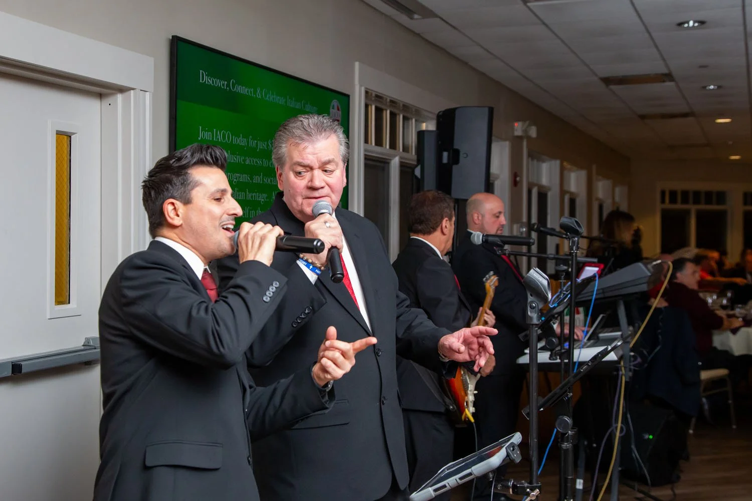 A group of five men singing and playing instruments at an event, two men in front are holding microphones, one is singing and pointing, the other is talking. The others are playing guitars and keyboards in the background.