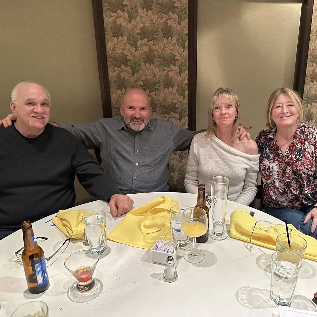 Four adults sitting at a table in a restaurant, smiling and posing for the photo. The table has drinks, yellow napkins, and a small box of sugar packets.