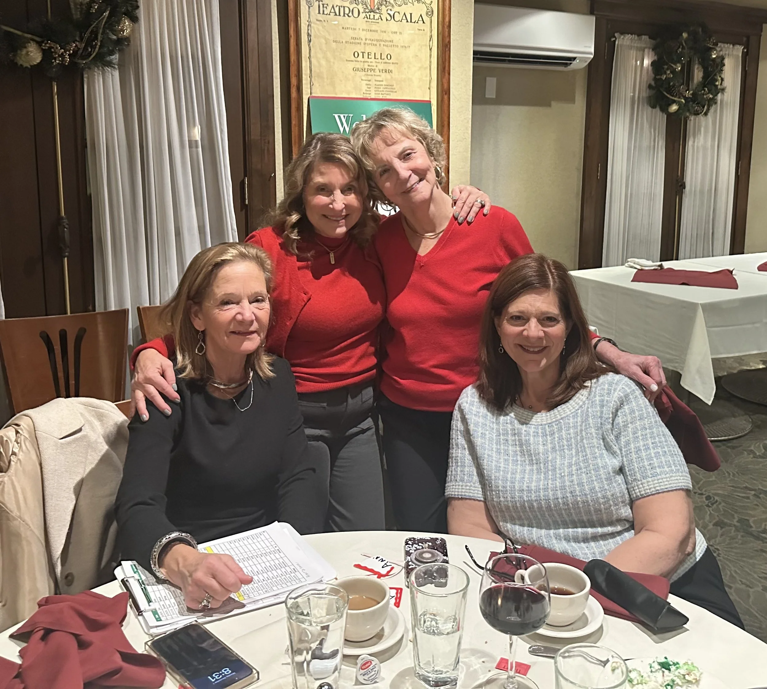 Four women gathered at a dining table for a holiday celebration, with two women standing behind and two seated in front, all smiling. The table has drinks, notebooks, and festive decorations.