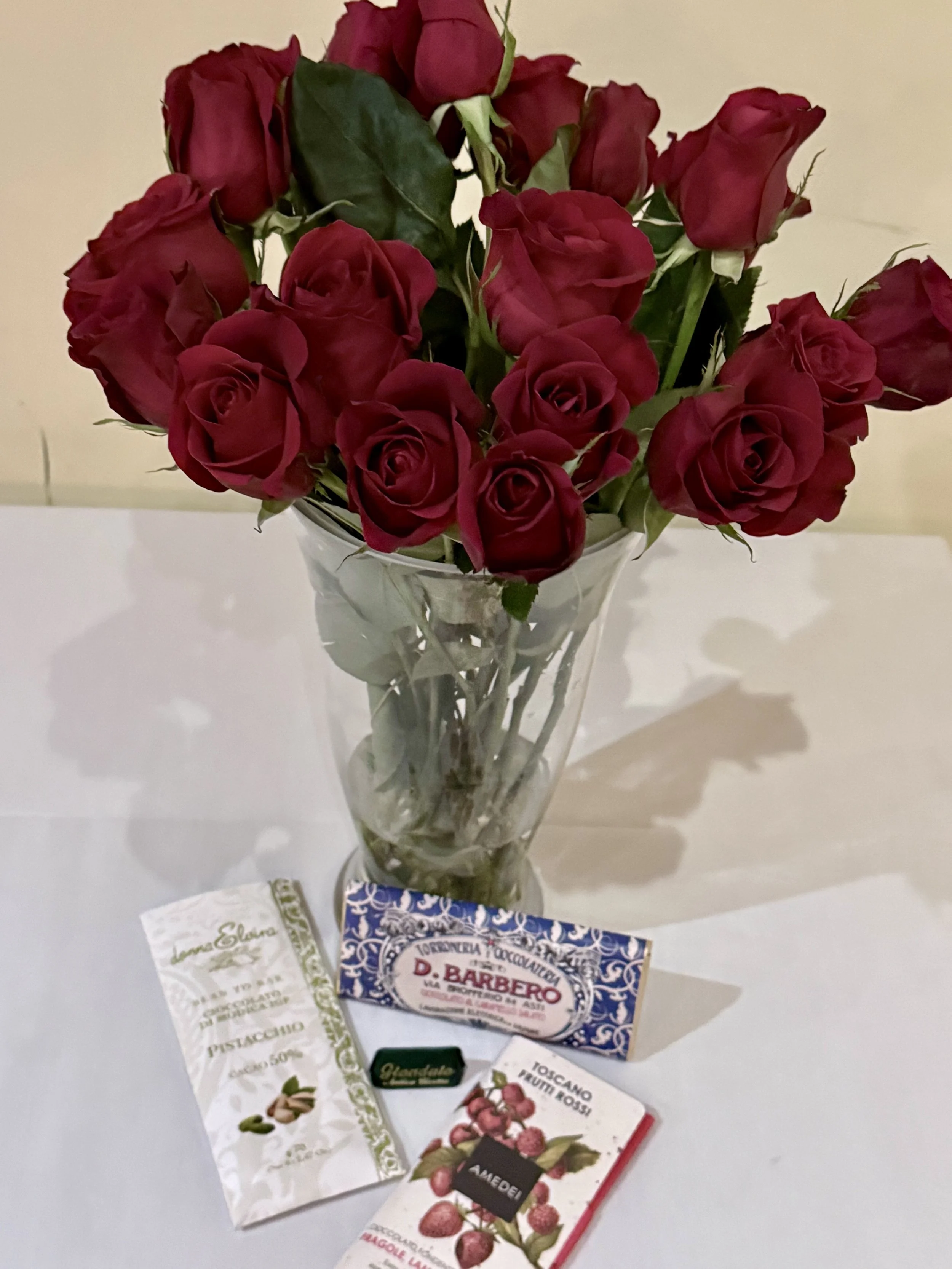A bouquet of red roses in a glass vase placed on a white surface, with various seed packets and a chocolate bar in front of it.