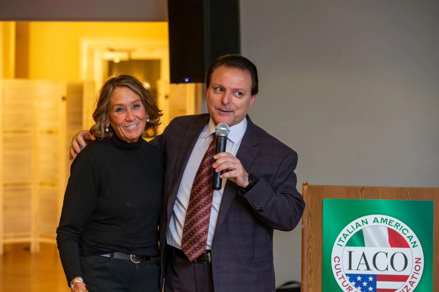 A woman and a man standing next to each other at a formal event. The woman is wearing a black turtleneck and the man is holding a microphone, wearing a suit and tie. There is a green and white logo on a wooden stand that reads 'IACO' and 'Italian Ame