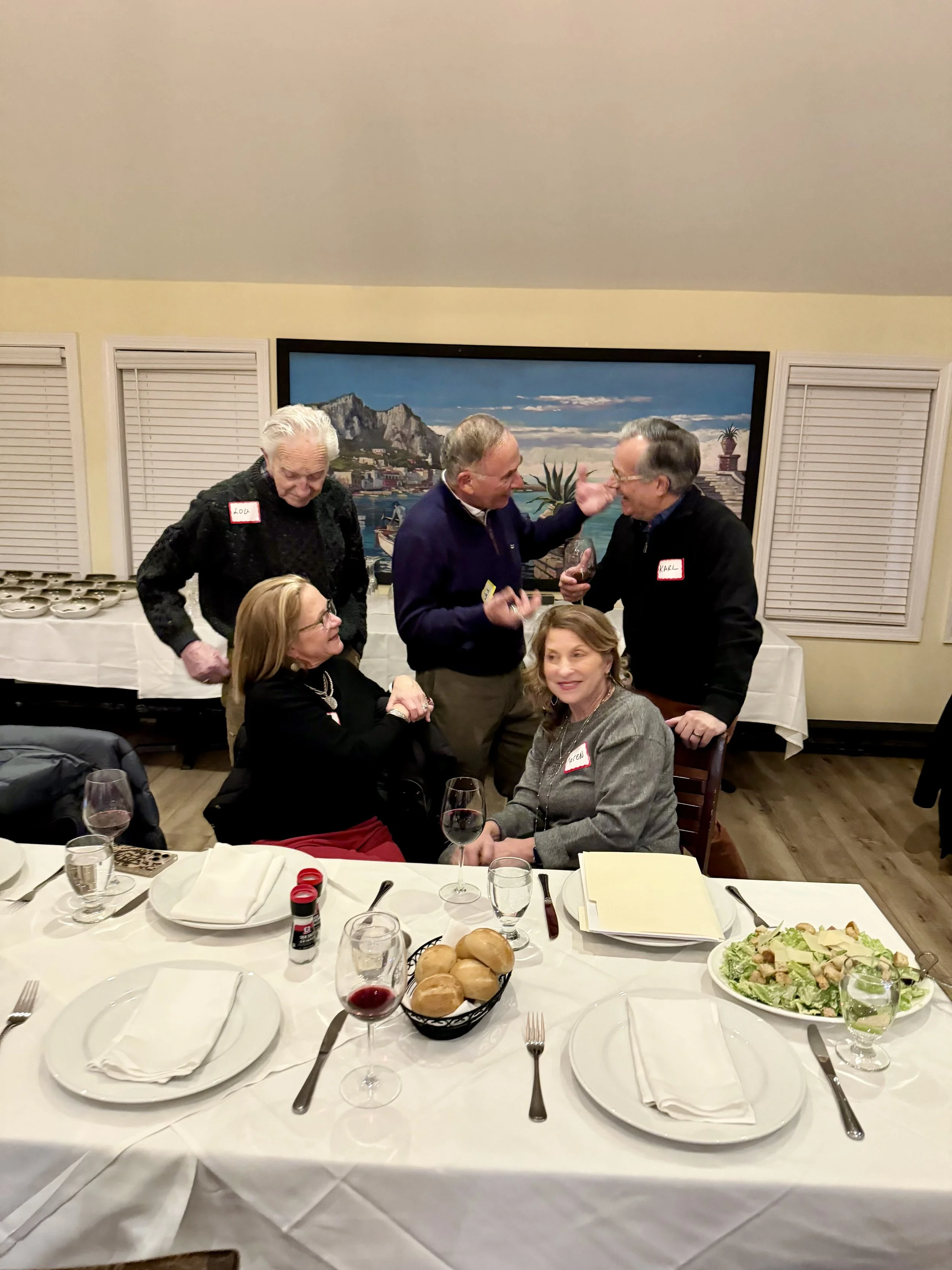 Group of five adults at a dinner party, with three men standing and two women seated, engaging in conversation and smiling, with a buffet table in the background and a painting on the wall.