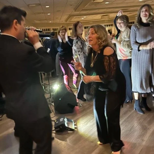 Woman holding an award and talking to a man with a microphone at an indoor event with a group of women in the background.