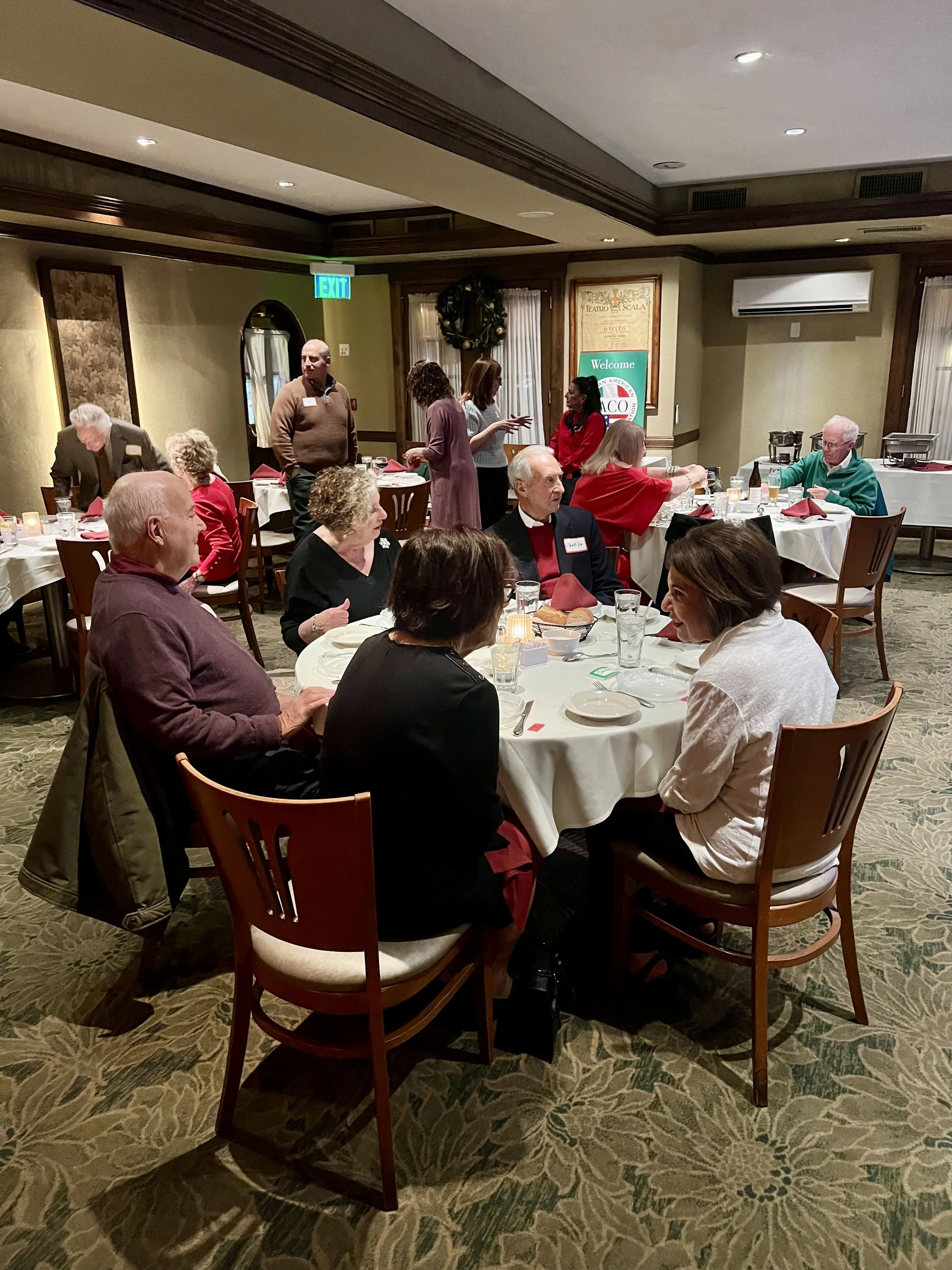 People sitting and talking at round tables in a restaurant or banquet hall decorated for a holiday gathering, with a sign in the background reading "Welcome" and a small stage or buffet area.
