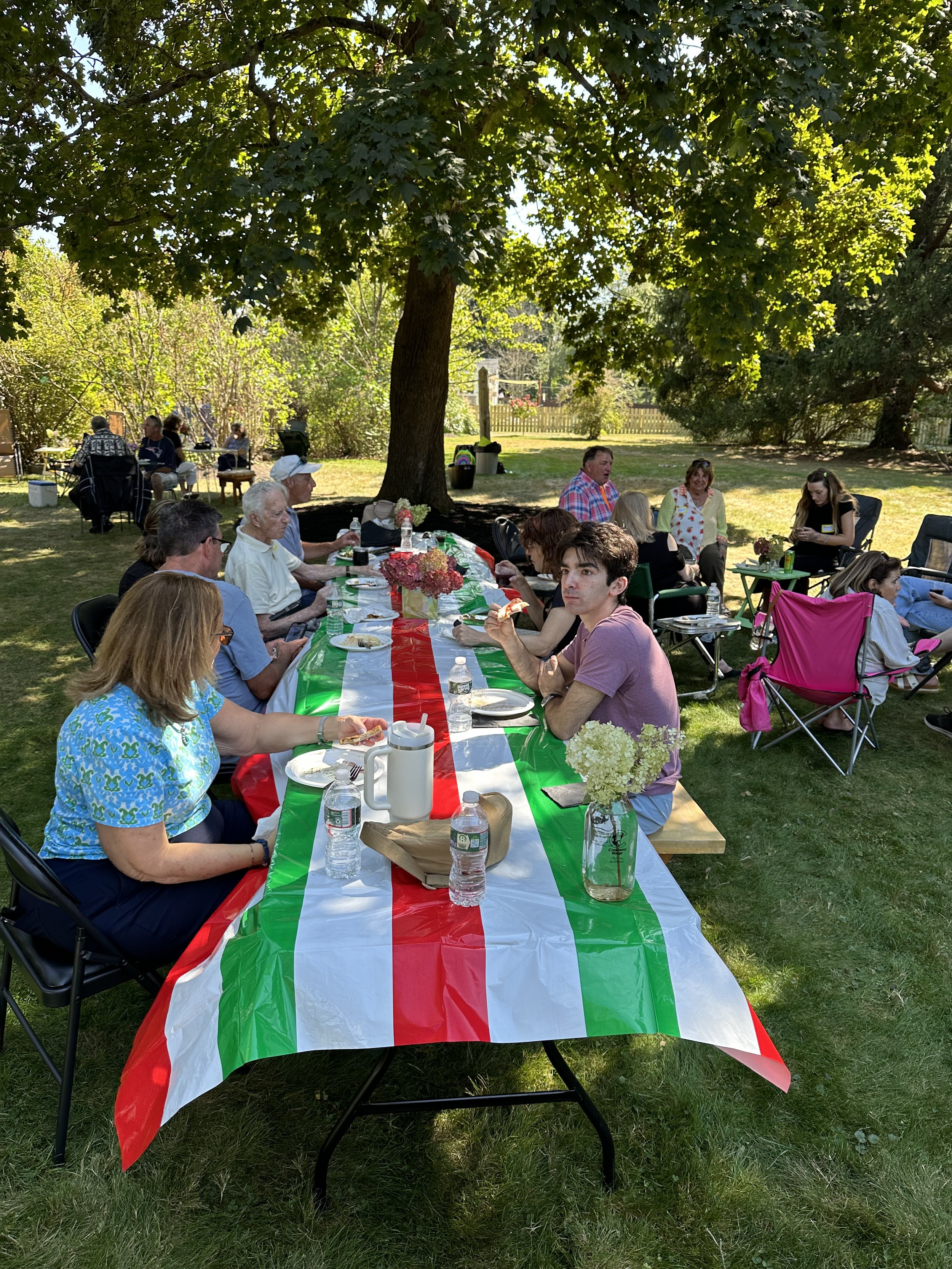 People gathered outdoors at a picnic table covered with a red, white, and green striped tablecloth, under a large tree. The group appears to be enjoying a meal together on a sunny day.