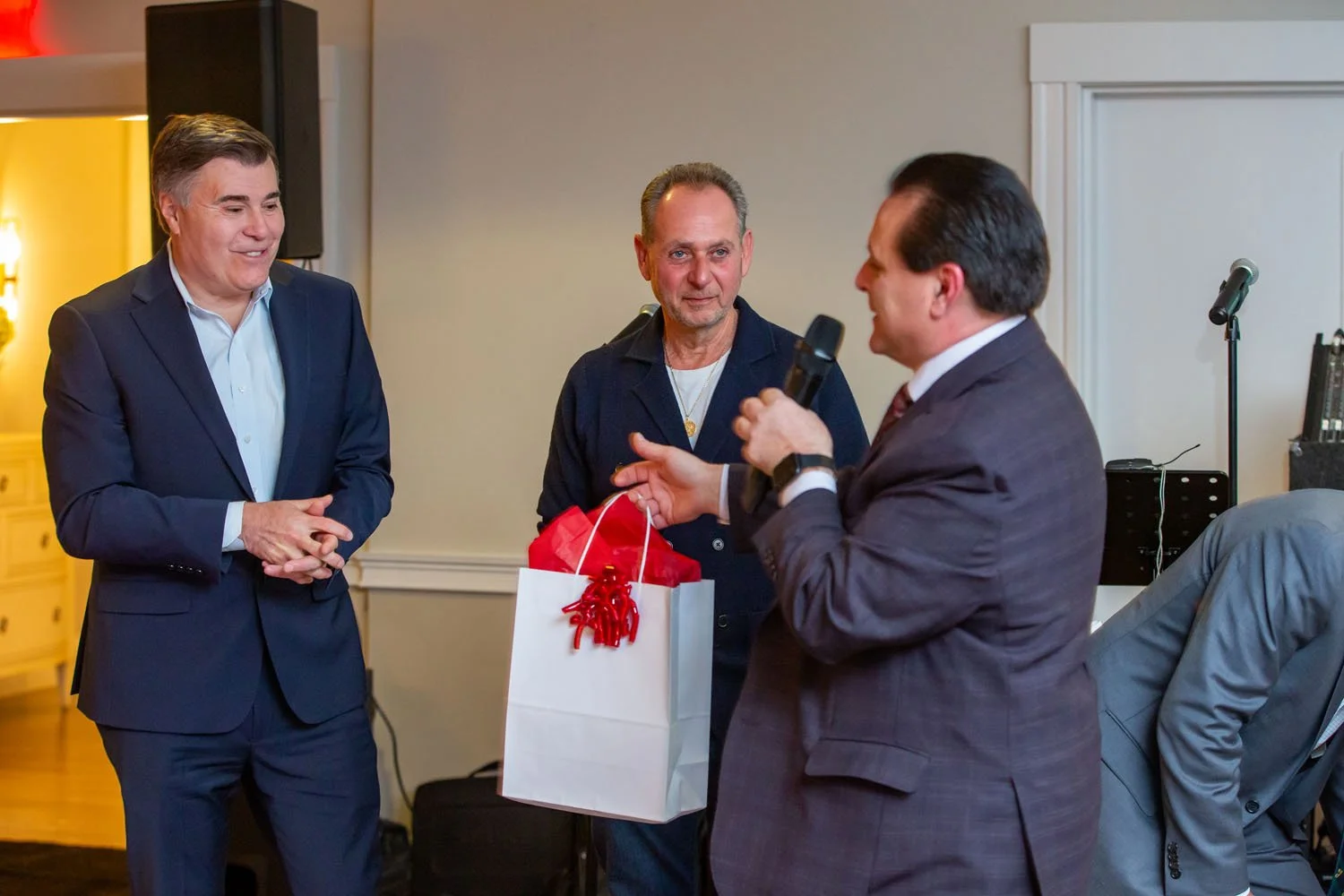 Three men at an indoor event, with one man in a suit speaking into a microphone, and the other two men listening; one of the listening men is holding a gift bag with a red ribbon.