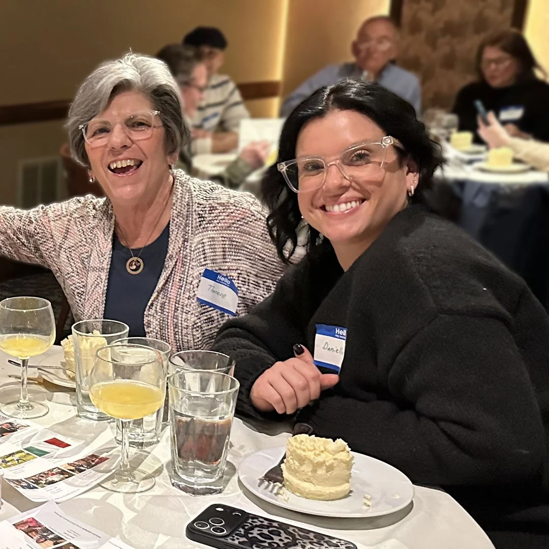 Two women smiling and sitting at a table with drinks and a cake slice, in a social gathering or event setting, with other people in the background.