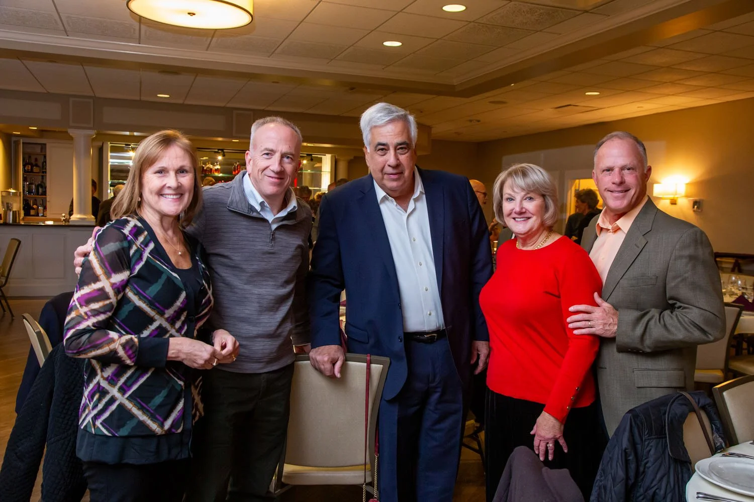 Group of five adults at a social gathering, standing together in a restaurant or banquet hall with warm lighting and visible bar area in the background.