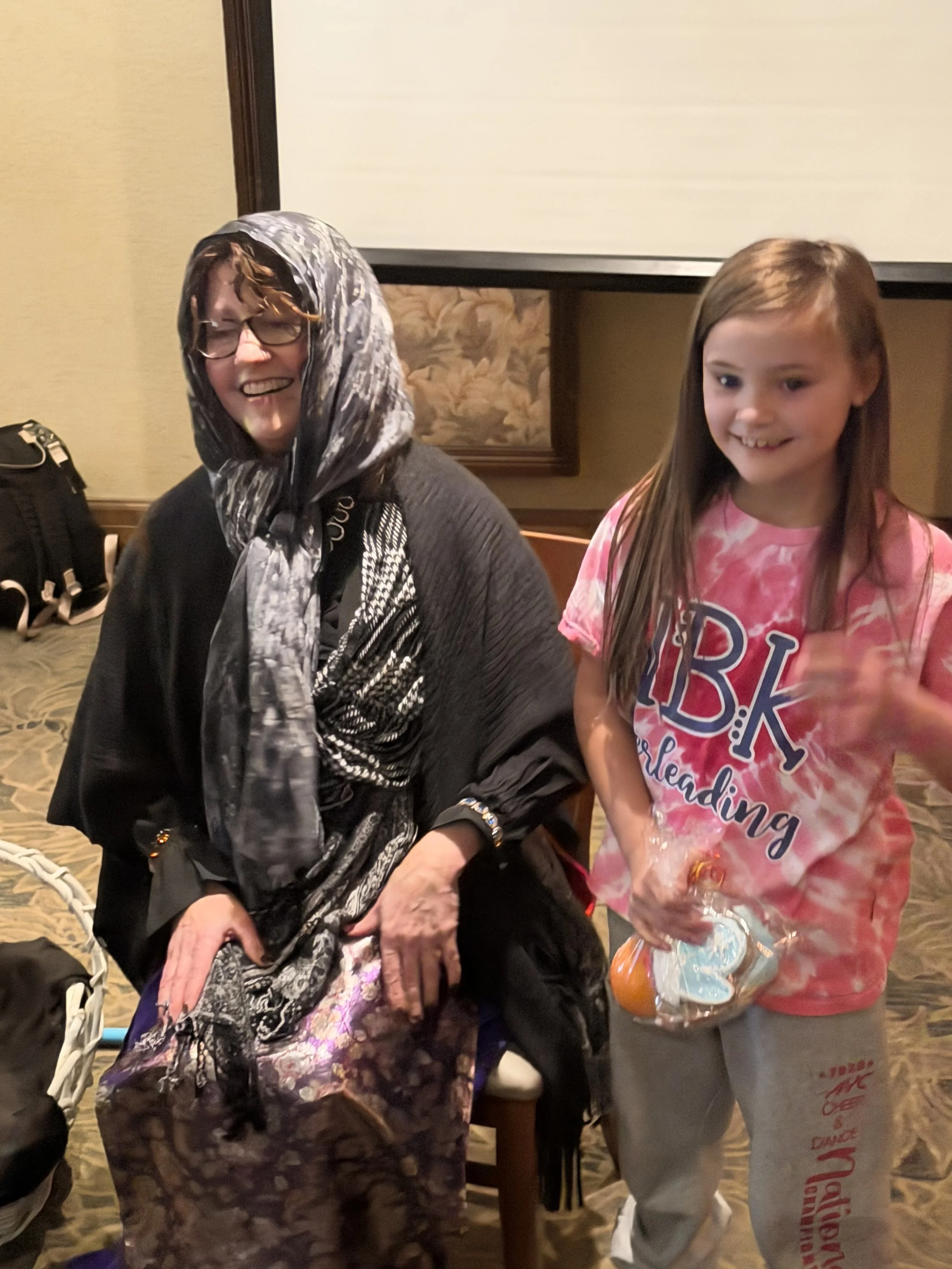 A woman wearing a headscarf and glasses sitting next to a girl in a pink tie-dye T-shirt with the words 'BKS Cheerleading' on it. The girl is holding a bag of marshmallows.