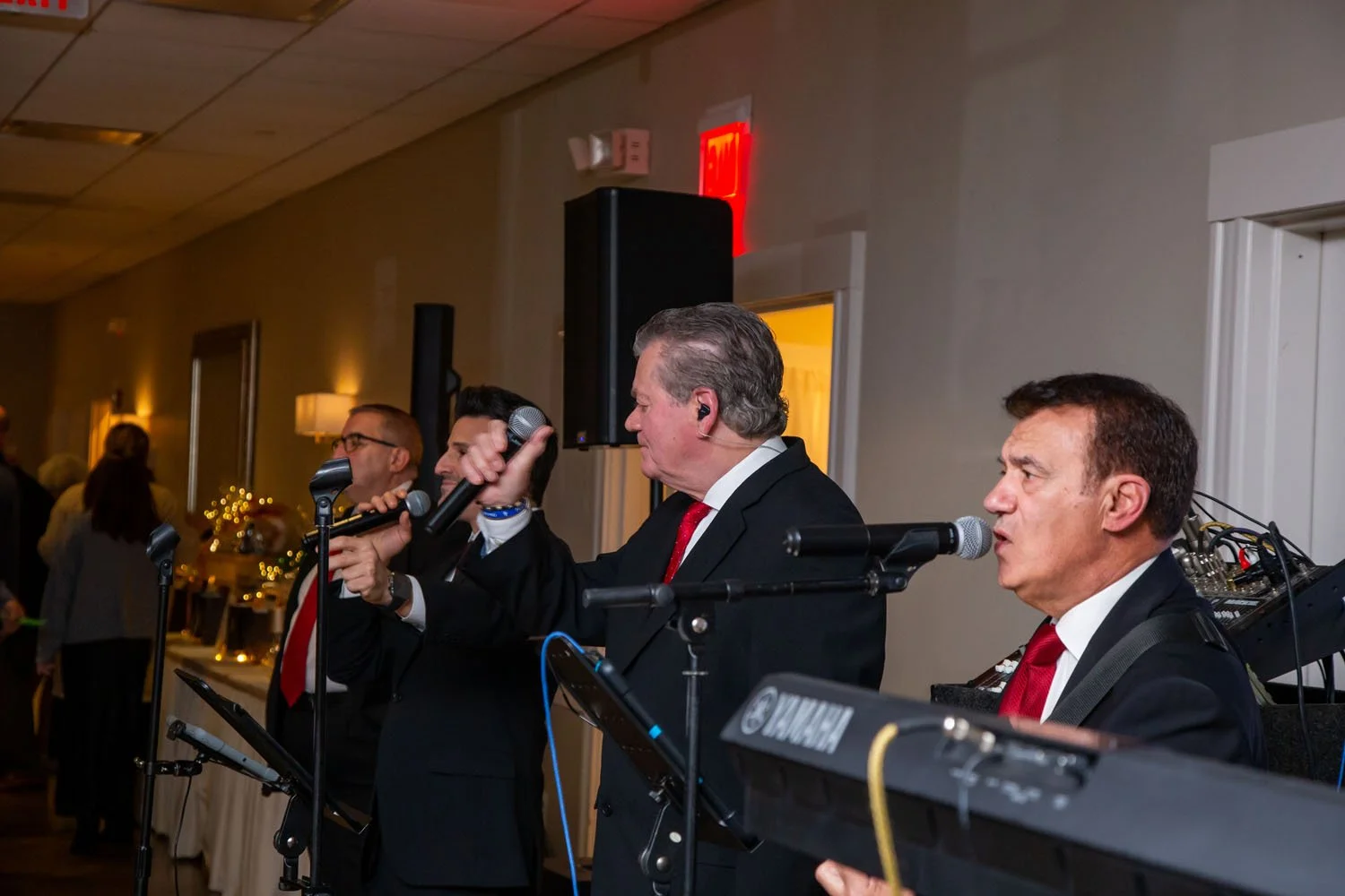 Three men dressed in black suits, white shirts, and red ties performing with microphones and musical instruments, likely at an indoor event or party.