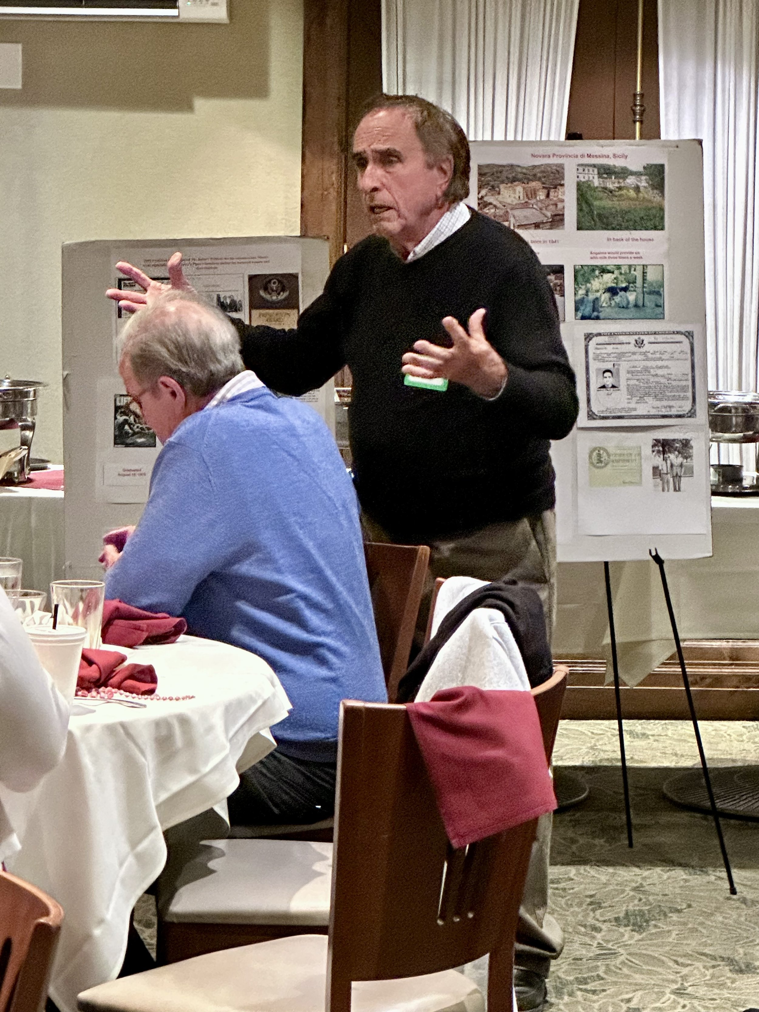 A man standing and speaking at an indoor event, with another person seated at a table listening. Background features display boards with photos and documents.
