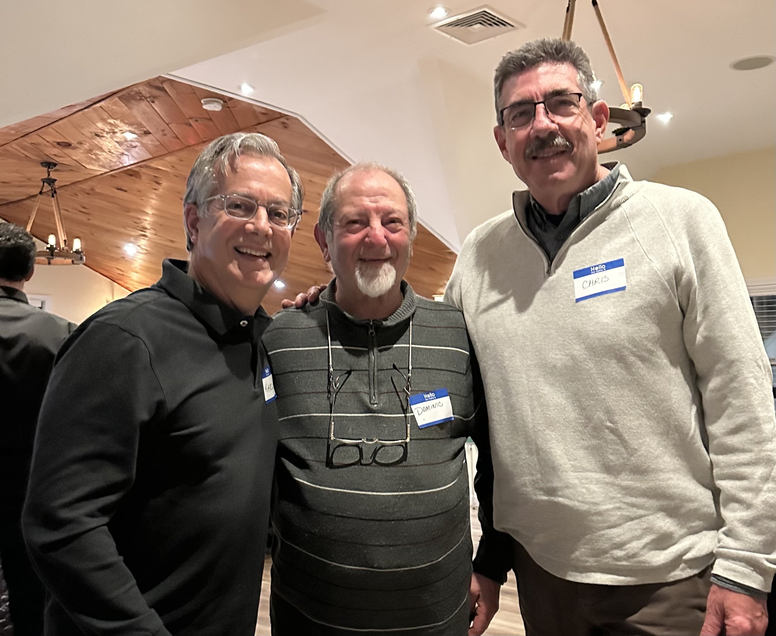 Three men smiling and posing indoors at an event, wearing name tags. The ceiling has wooden panels and a hanging light fixture.