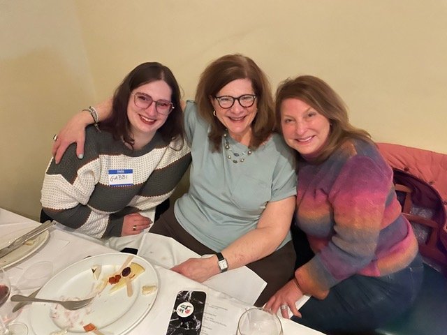 Three women smiling and seated at a dining table, with plates and glasses in front of them.