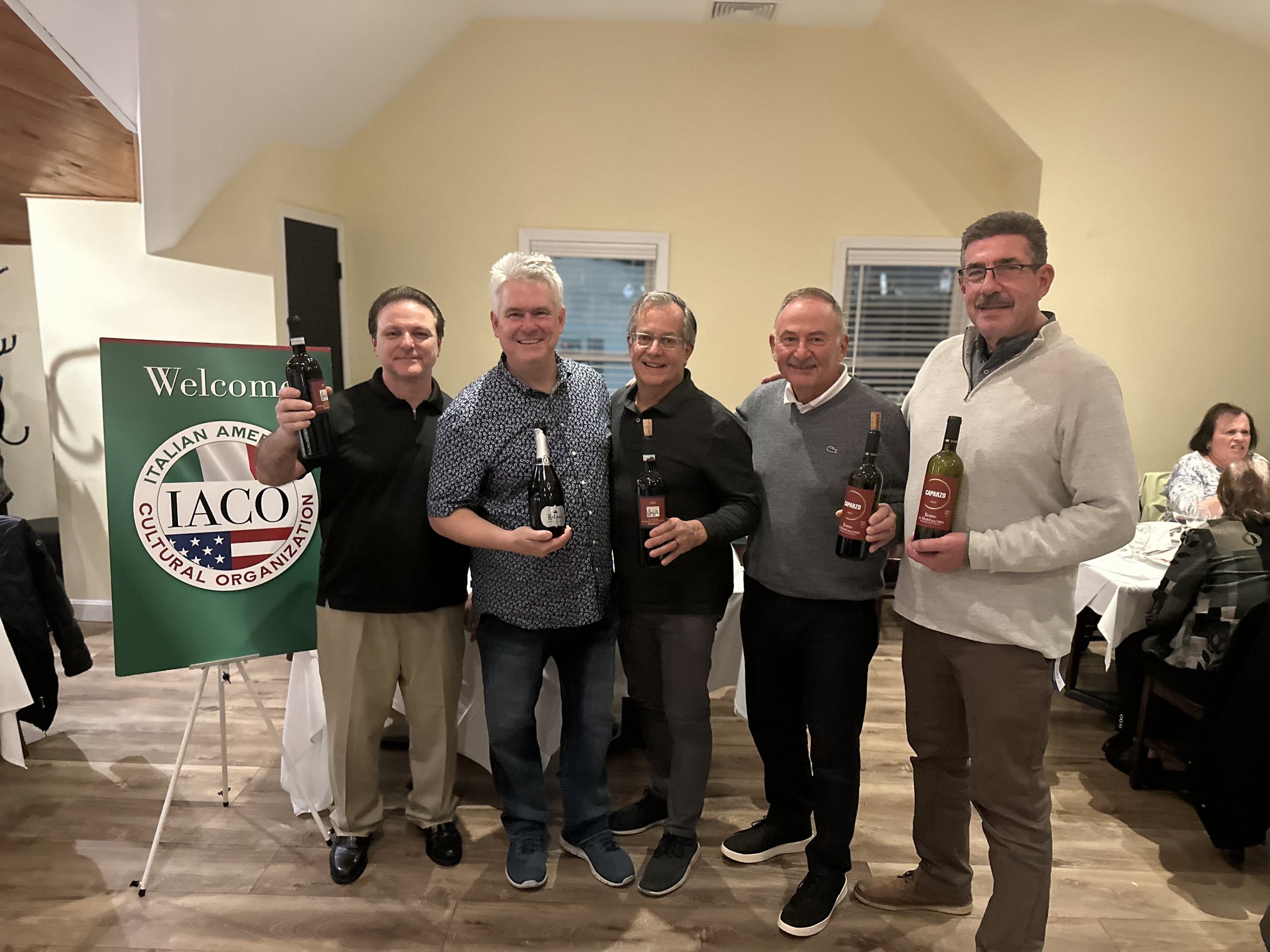 Five men holding wine bottles stand in front of a sign for the Italian American Cultural Organization at an indoor event.