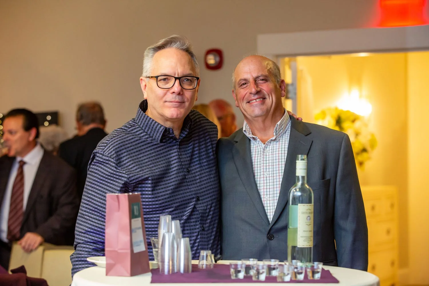 Two men in formal attire standing together at a social gathering, smiling with drinks and bottles on the table in front of them.