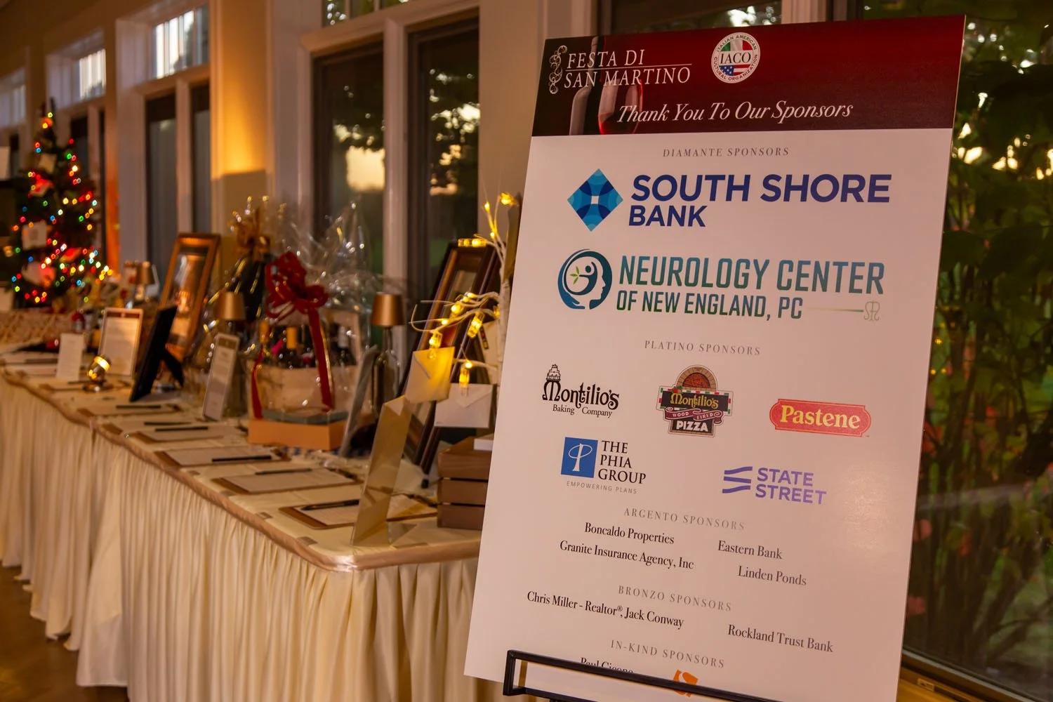 Large signboard listing sponsors for the Festa di San Martino event, with a decorated table in the background that holds gifts and framed photos. The background shows windows and a Christmas tree with colorful lights.