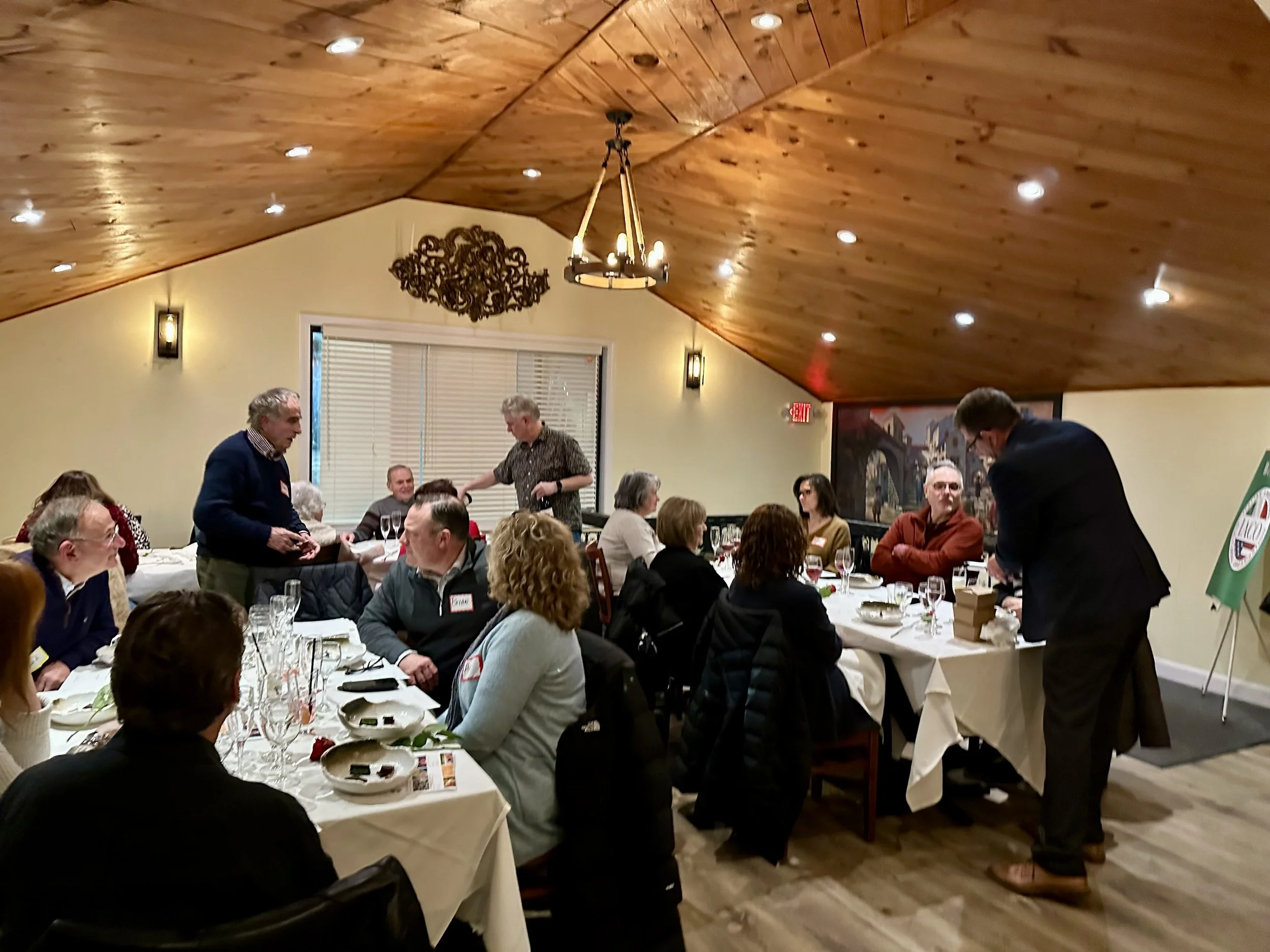 People gathered in a restaurant for a social event, sitting around tables with plates, glasses, and silverware, while some are engaging in conversation or being served food, with warm wooden ceiling and decorative lighting.
