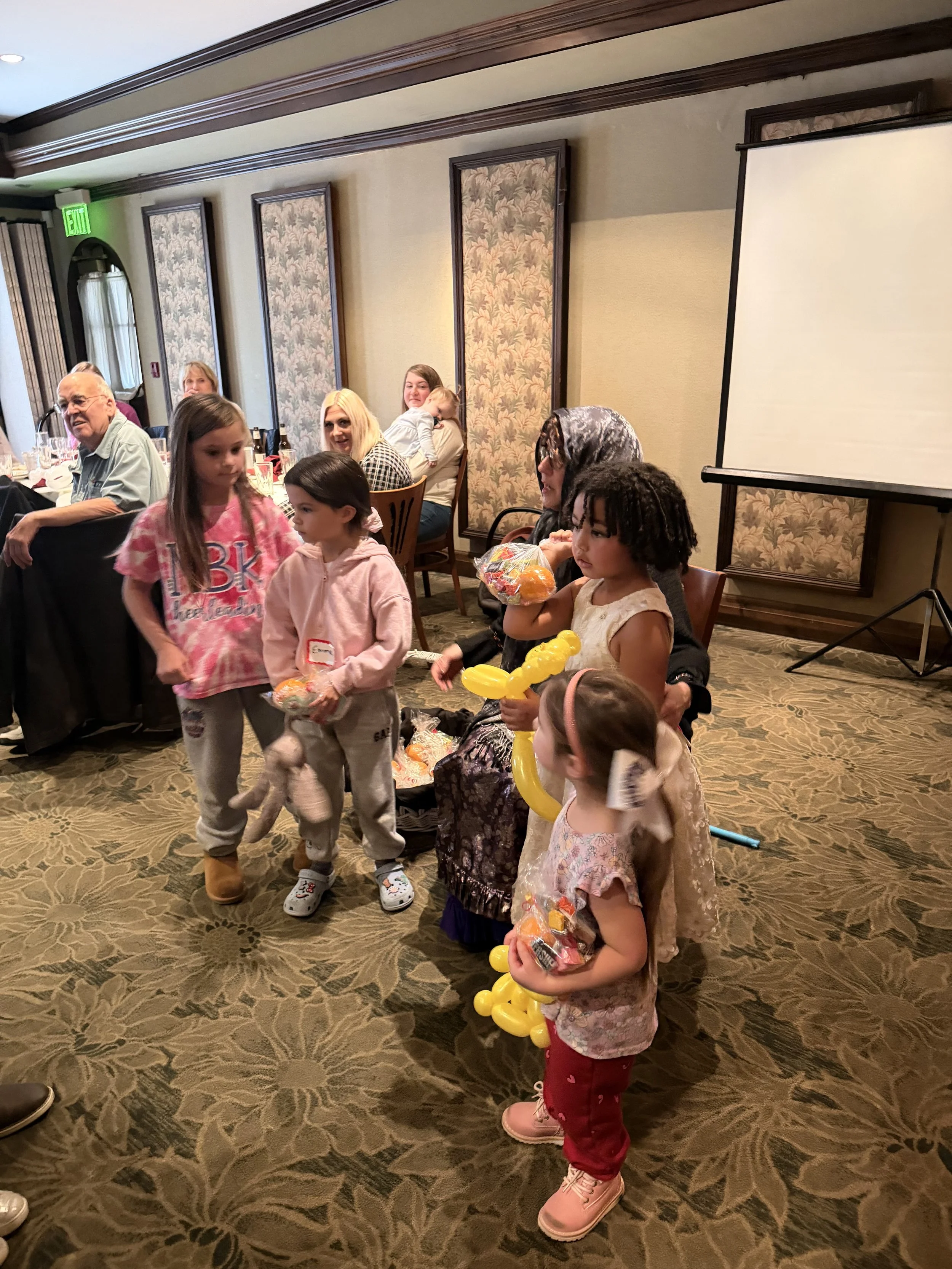 Children receiving small toy animals and balloon animals at a party, while adults are seated at a long table in the background in a decorated banquet room.