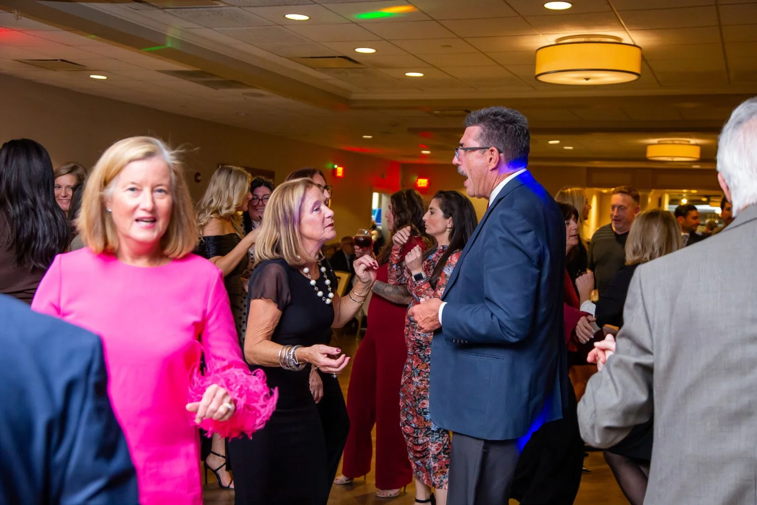 People gathered and socializing at an indoor event or party. Some are holding drinks, and the room is decorated with warm lighting.